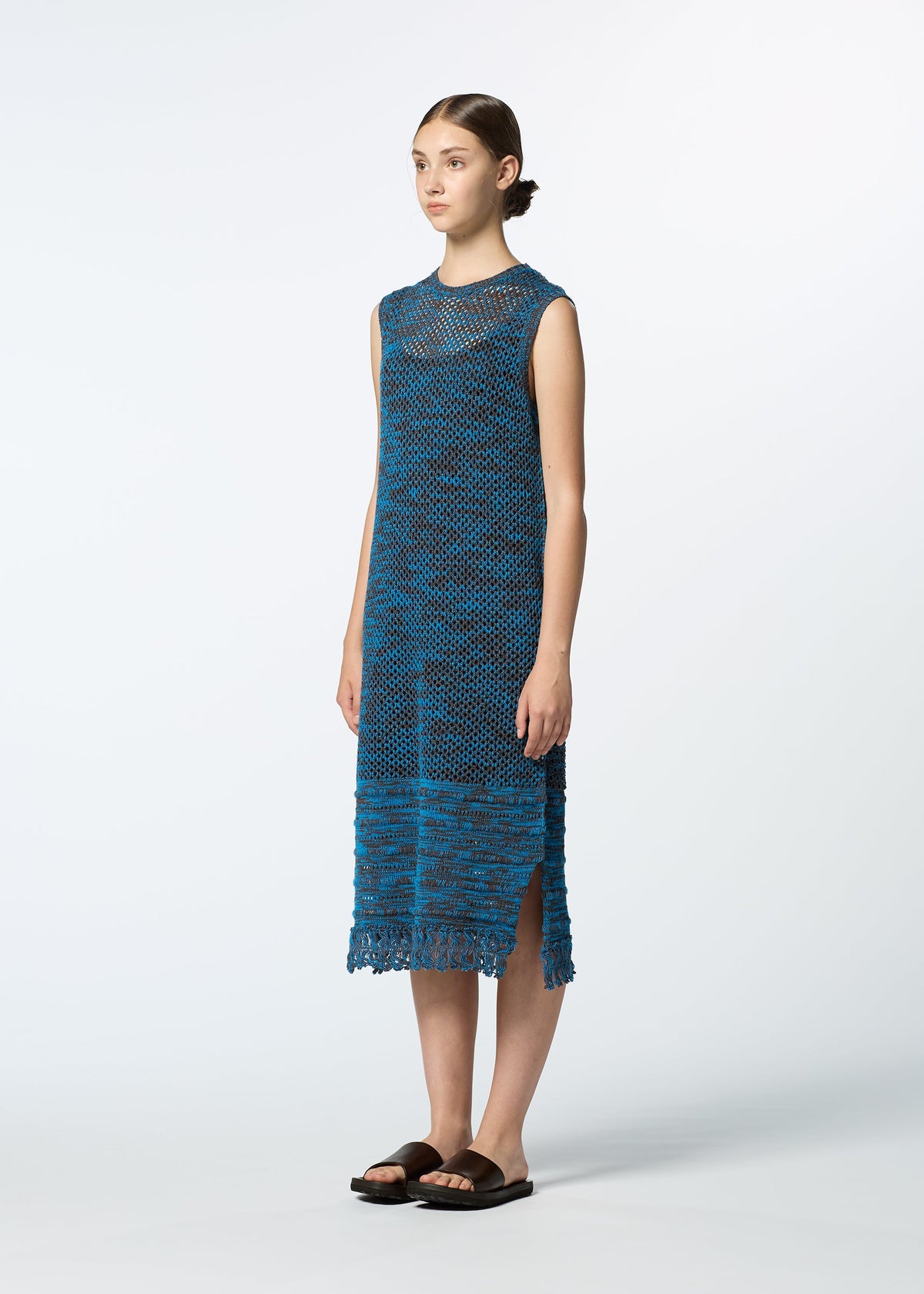 A woman stands in profile wearing HaaT's MELANGE MESH sleeveless, blue knit dress with side slits and black sandals. Her hair is tied back in a low bun against a plain white background. view 5 of 6