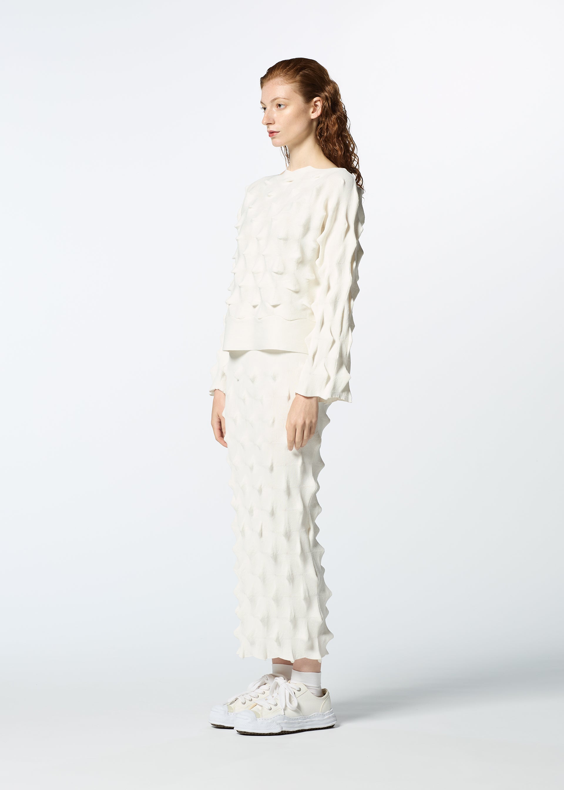 Zoomed view A person with long, wavy hair stands sideways in HaaT’s ZIG ZAG KNIT top and skirt, made from organic cotton and recycled polyester, paired with white sneakers against a light background.