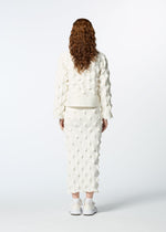 A person with long, curly brown hair faces away, wearing HaaT's ZIG ZAG KNIT white textured organic cotton top and matching skirt set with white sneakers, set against a plain white background.