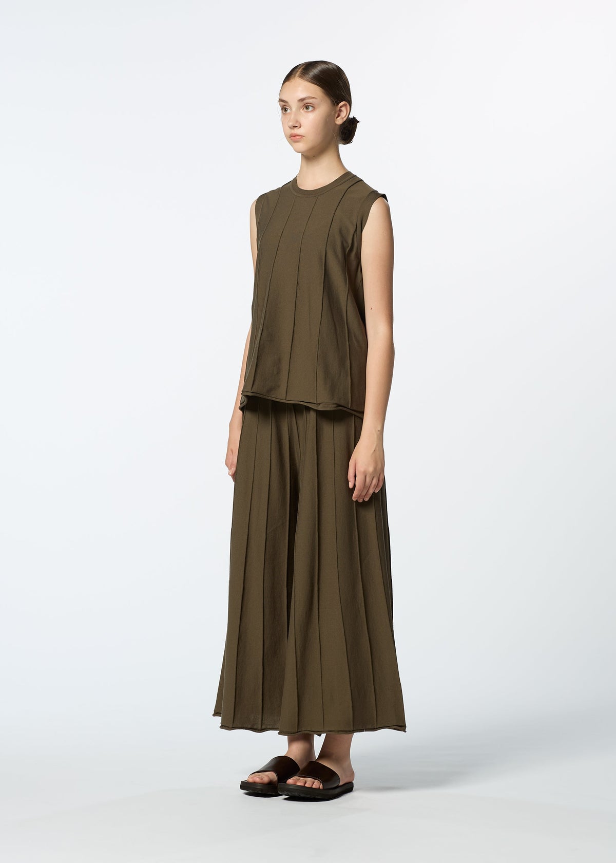 A woman stands in a studio against a plain white background, wearing HaaT's RADIAL LINE KNIT—an olive-green, sleeveless, loose top and matching wide-leg skirt in recycled polyester—with her hair pulled back and black slide sandals. view 7 of 8