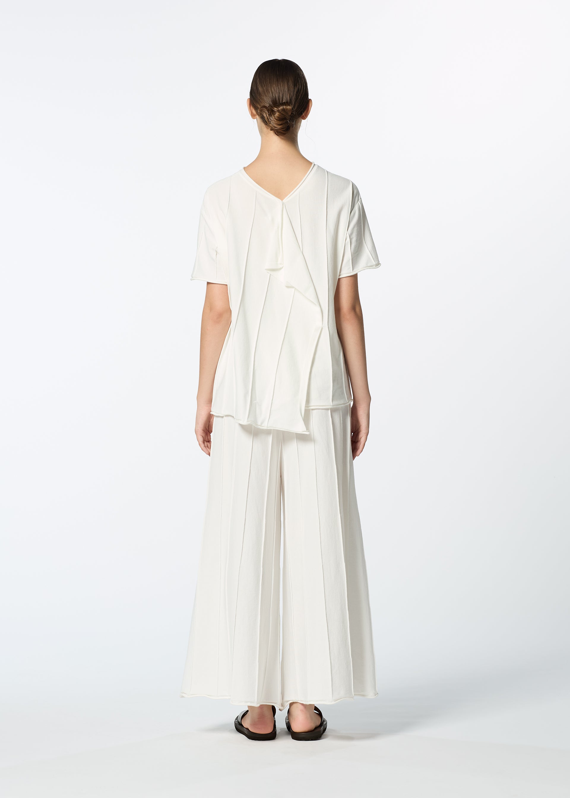 Zoomed view A person stands facing away, wearing HaaT's RADIAL LINE KNIT—a loose-fitting short-sleeved top with a vertical seam and ruffle detail at the back—paired with wide-leg white pants and black flats, set against a plain white background.