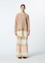A person faces forward against a plain white background, wearing HaaT's MELANGE MESH loose, textured organic cotton sweater with wide-legged, color-blocked pants in cream, tan, and pale pink tones, paired with black sandals.
