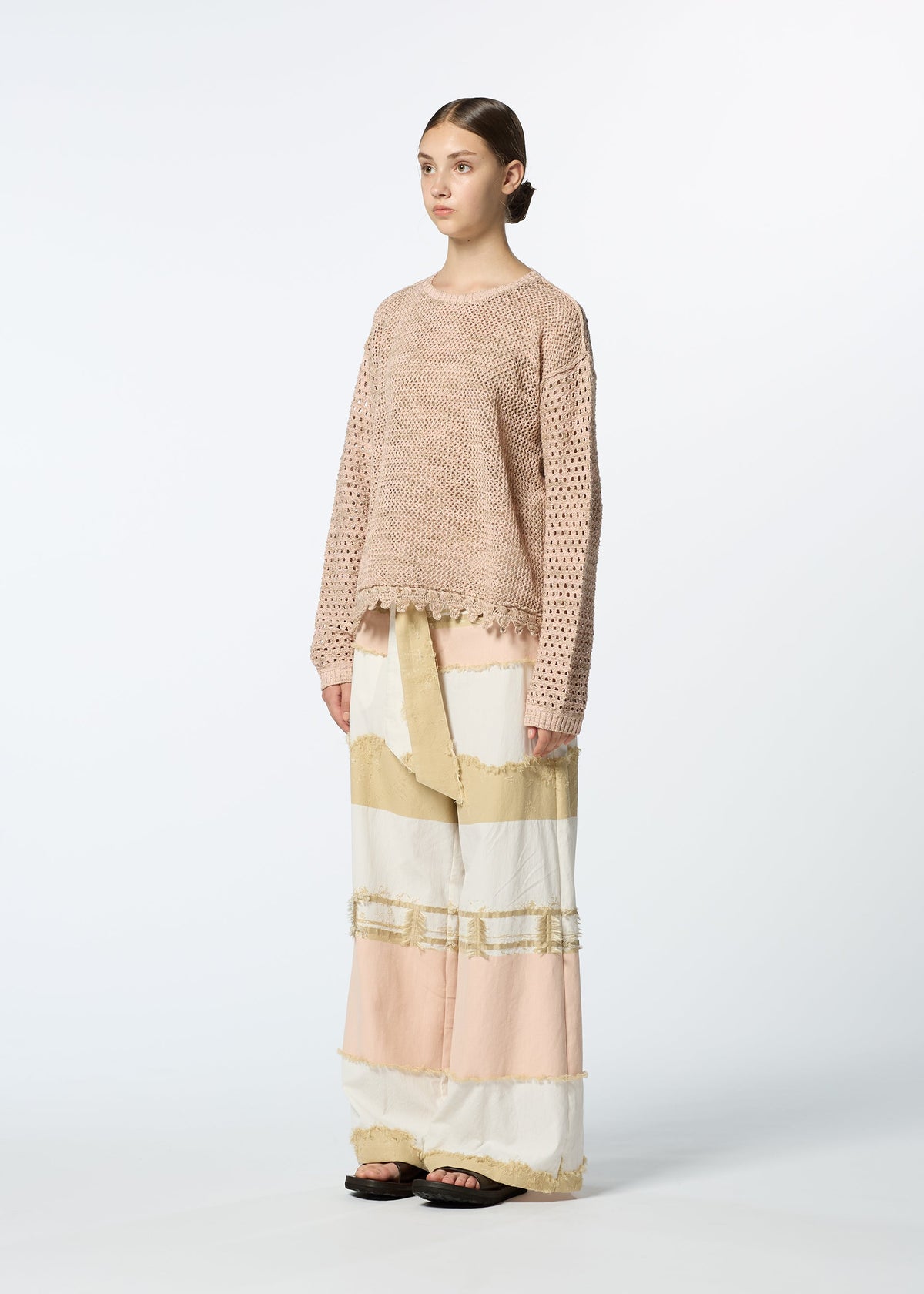 A person stands against a plain background wearing HaaT's MELANGE MESH, a loose light pink organic cotton knit top, paired with a long color-blocked skirt in cream, beige, and blush tones and black sandals for a casual, relaxed look. view 5 of 7