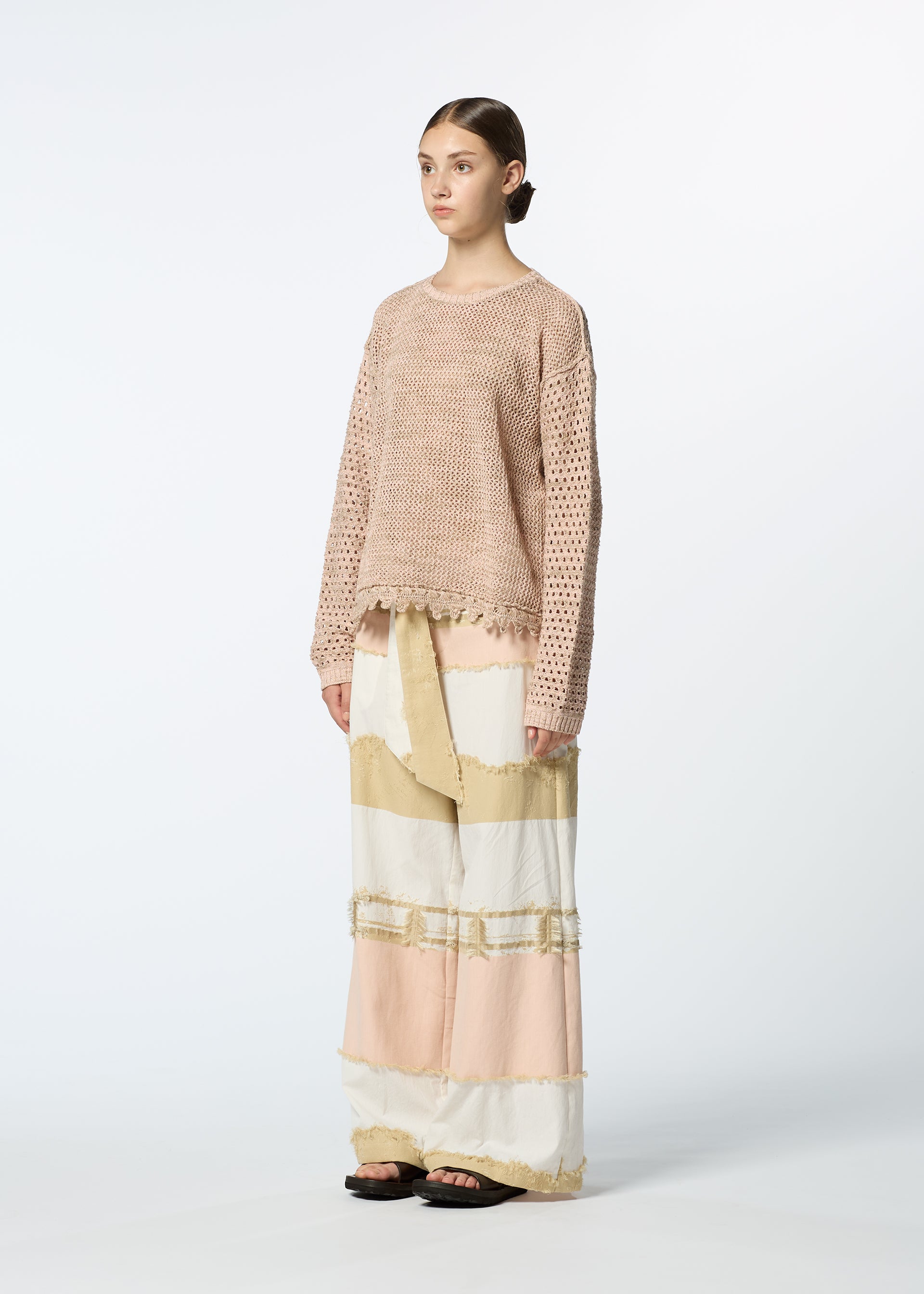 Zoomed view A person stands against a plain background wearing HaaT's MELANGE MESH, a loose light pink organic cotton knit top, paired with a long color-blocked skirt in cream, beige, and blush tones and black sandals for a casual, relaxed look.