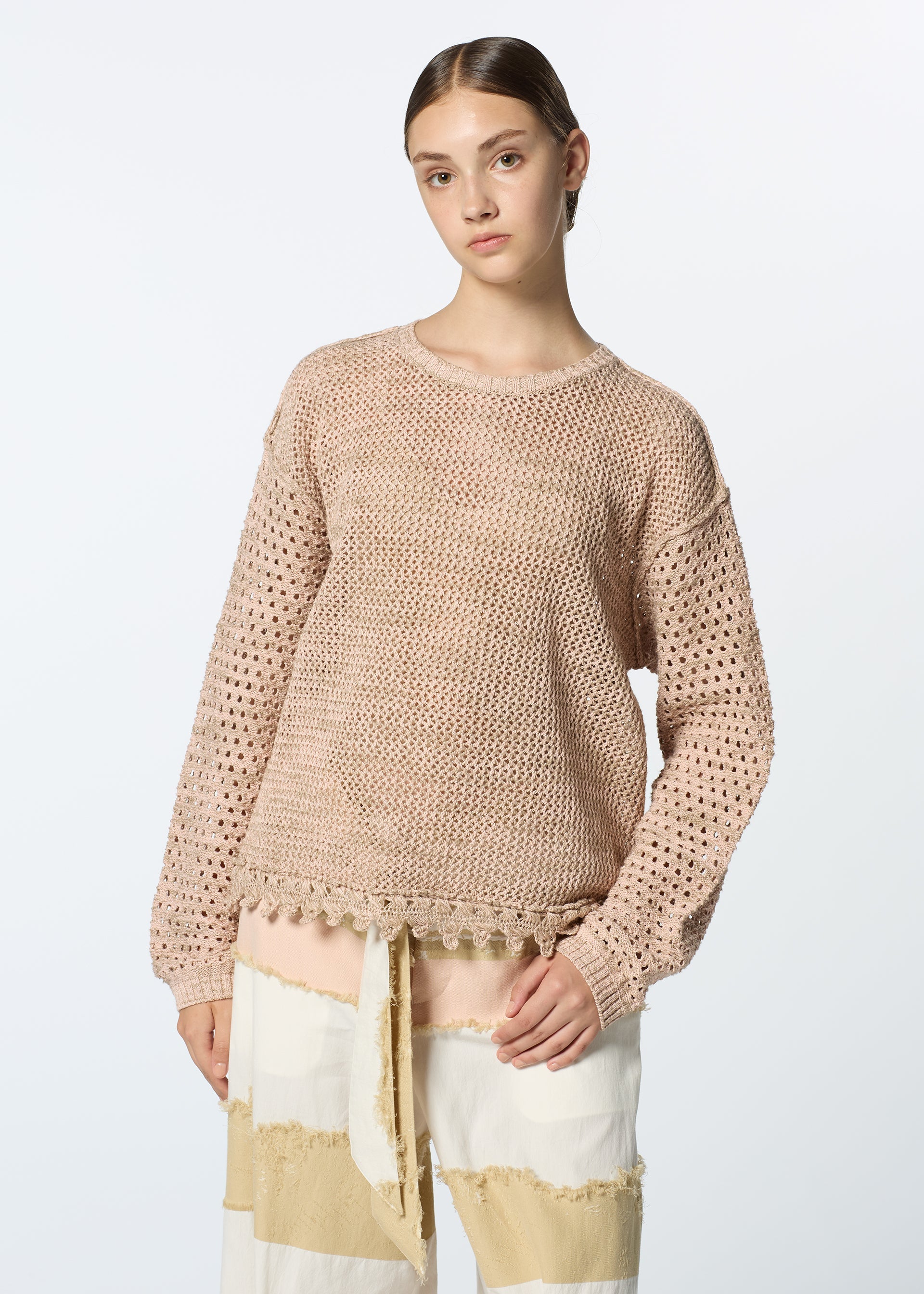 Zoomed view A young woman with straight brown hair models HaaT’s MELANGE MESH: a loose-knit, light pink top with crochet sleeves and mesh lace hem, paired with wide-leg, cream and tan patchwork pants against a plain light background.