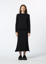 A person with long, curly red hair faces forward, wearing HaaT's PLATED LINES set—a plain black lightweight knit top and matching ankle-length skirt made from recycled polyester—paired with black sneakers, against a white background.