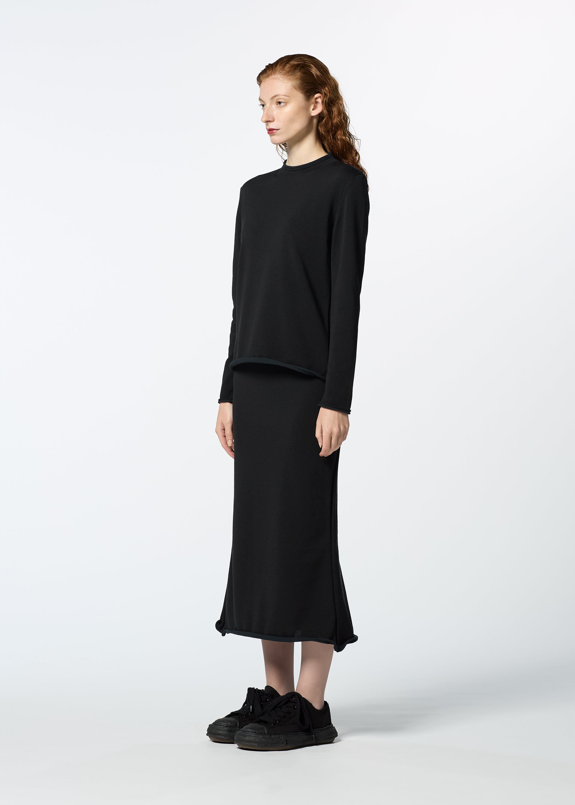 Zoomed view A person with long wavy hair stands sideways, wearing HaaT's PLATED LINES: a loose black lightweight knit top and matching ankle-length skirt made from recycled polyester, paired with black sneakers against a plain light background.