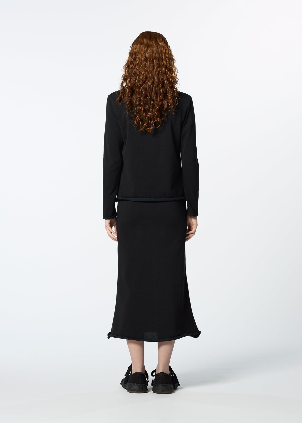 A person with long curly red hair, wearing HaaT's PLATED LINES black lightweight knit top and black midi skirt, stands facing away from the camera against a plain white background. view 8 of 8
