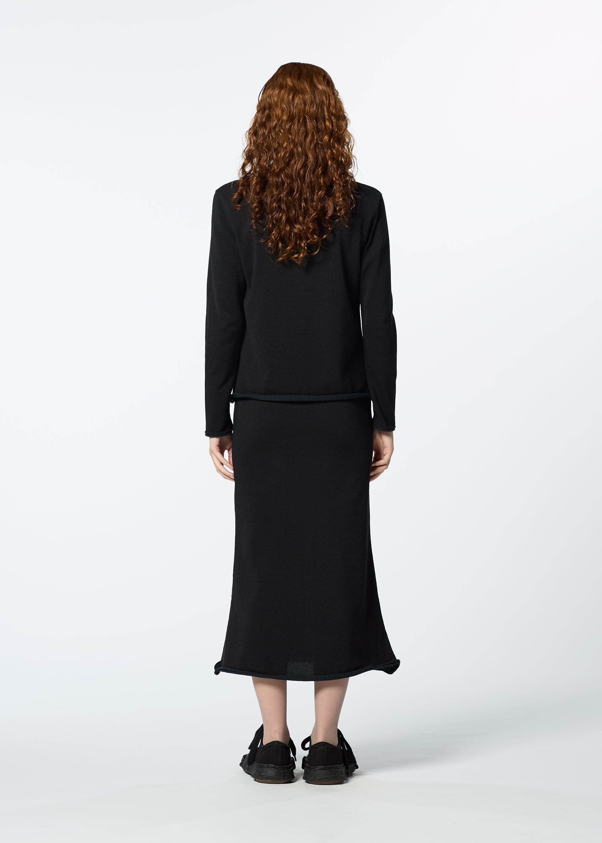 Zoomed view A person with long curly red hair, wearing HaaT's PLATED LINES black lightweight knit top and black midi skirt, stands facing away from the camera against a plain white background.