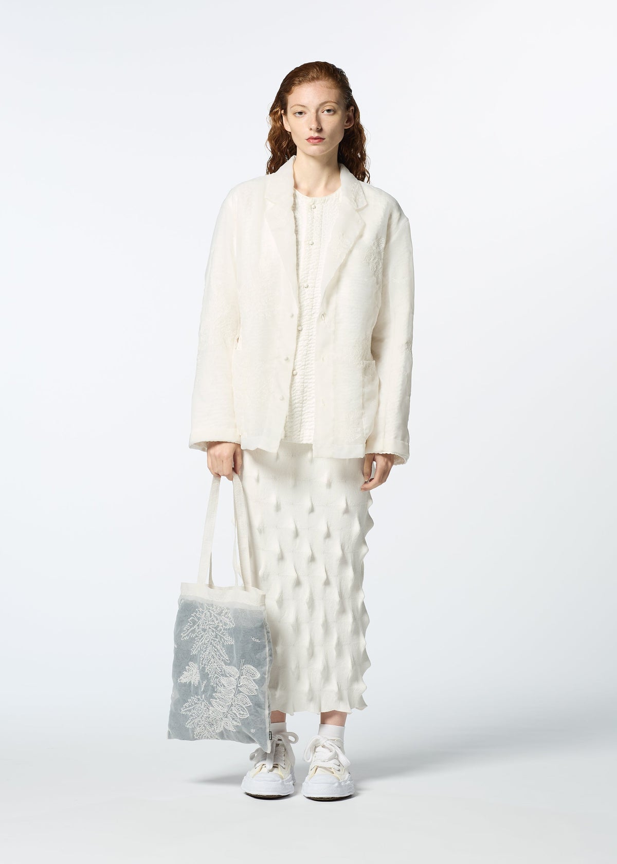 A woman with long, wavy, auburn hair stands facing forward in a textured white jacket and skirt with white sneakers, holding HaaT's GLASSLEAF TOTE JANUARY—a light-colored bag featuring a delicate leaf motif—against a plain background. view 7 of 8