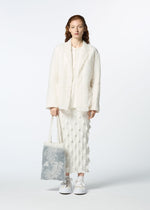 A woman with long, wavy, auburn hair stands facing forward in a textured white jacket and skirt with white sneakers, holding HaaT's GLASSLEAF TOTE JANUARY—a light-colored bag featuring a delicate leaf motif—against a plain background.
