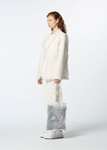 A person with wavy auburn hair stands against a plain light background, wearing an off-white textured suit and white sneakers, holding HaaT’s GLASSLEAF TOTE JANUARY—a sheer bag adorned with a delicate leaf motif.