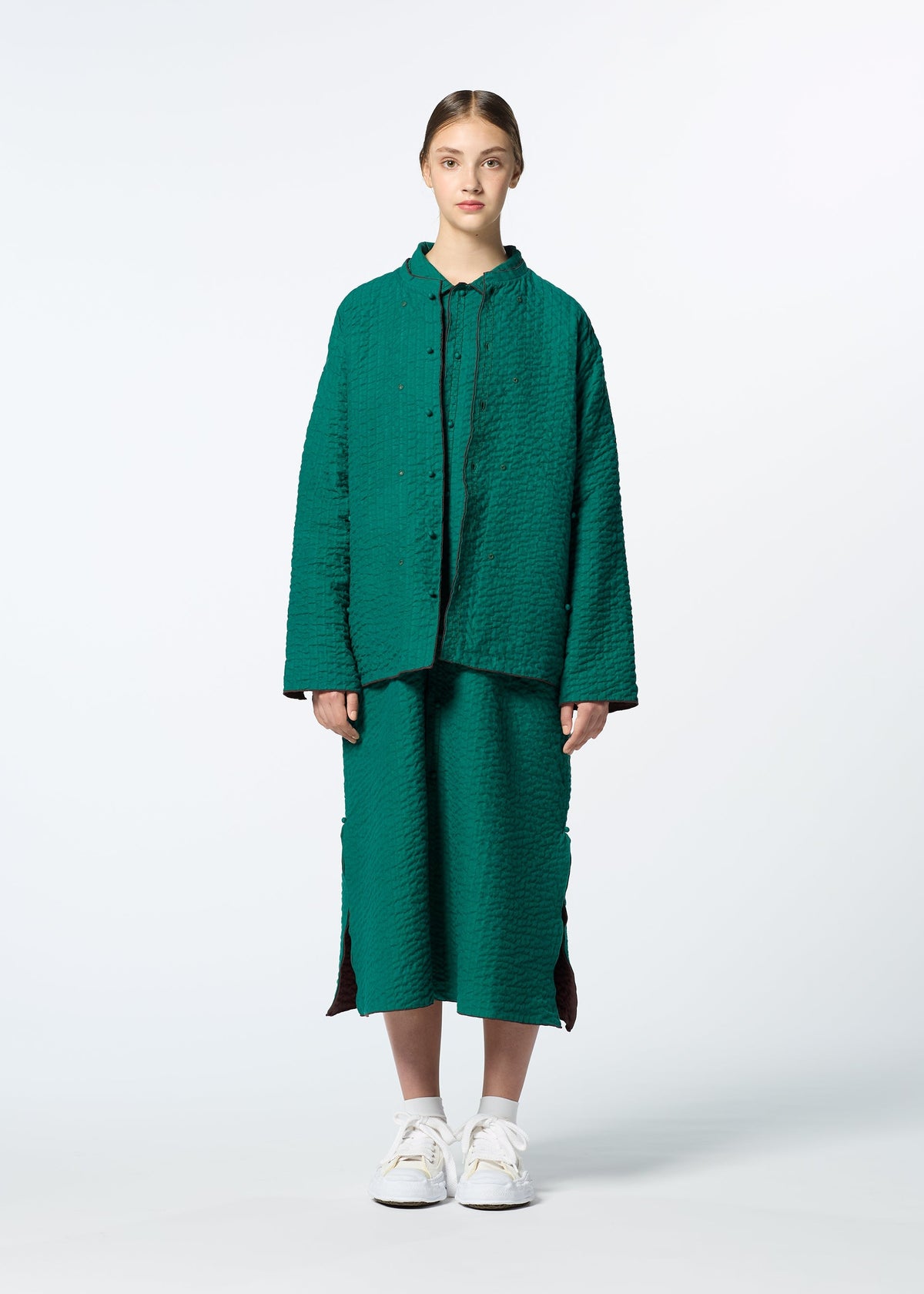 A young woman faces forward in HaaT’s DUAL LAYER set—a textured green button-up shirt and matching skirt with side slits, inspired by Kabira embroidery. She wears white sneakers, has her hair tied back, and stands against a plain light background. view 6 of 8