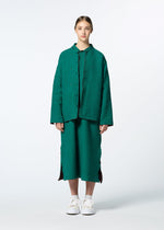 A young woman faces forward in HaaT’s DUAL LAYER set—a textured green button-up shirt and matching skirt with side slits, inspired by Kabira embroidery. She wears white sneakers, has her hair tied back, and stands against a plain light background.