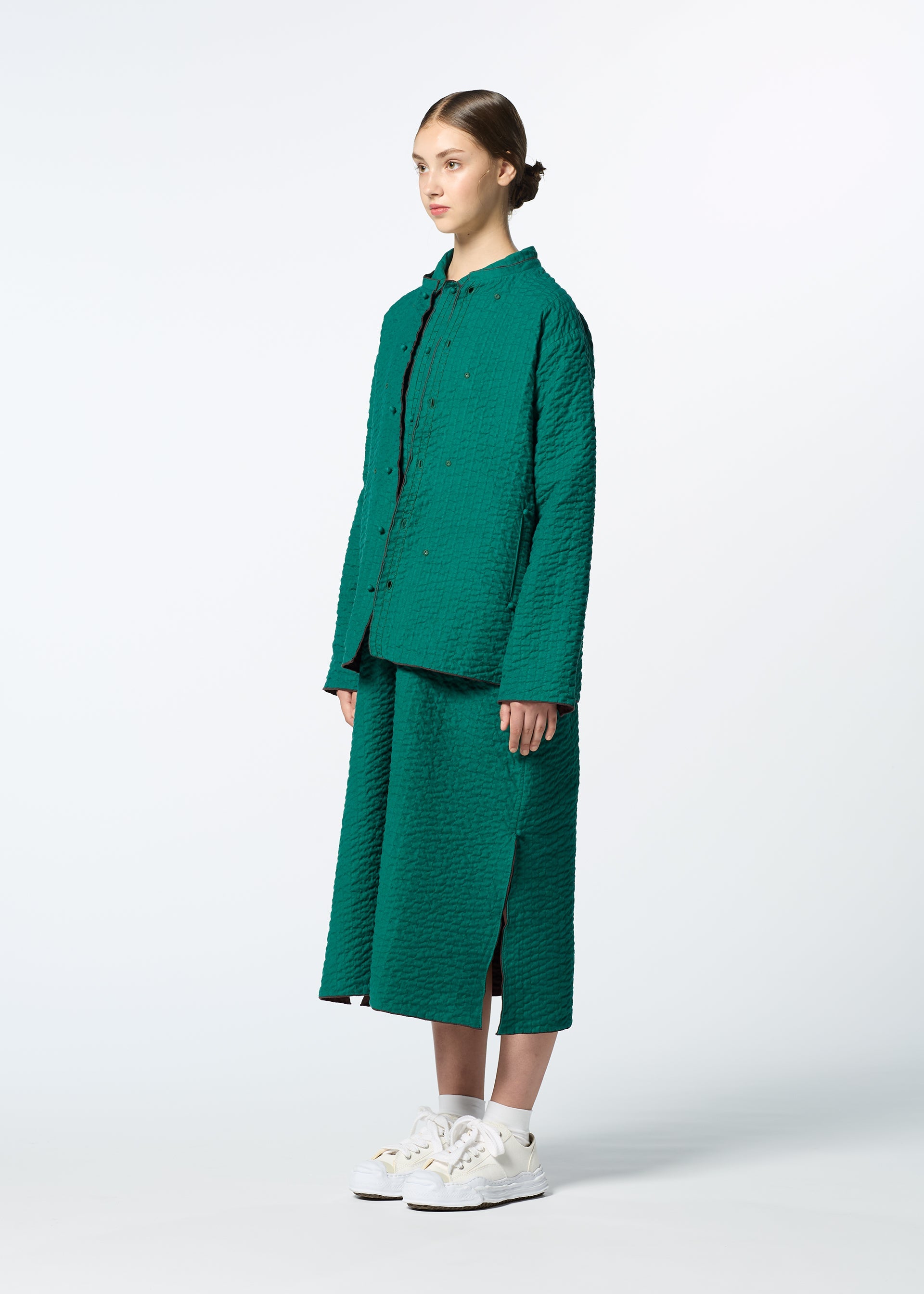 Zoomed view A person with brown hair tied back wears HaaT’s DUAL LAYER handwoven cotton jacket with Kabira embroidery and a matching teal midi skirt, paired with white sneakers—celebrating Indian artisanship against a plain light background.