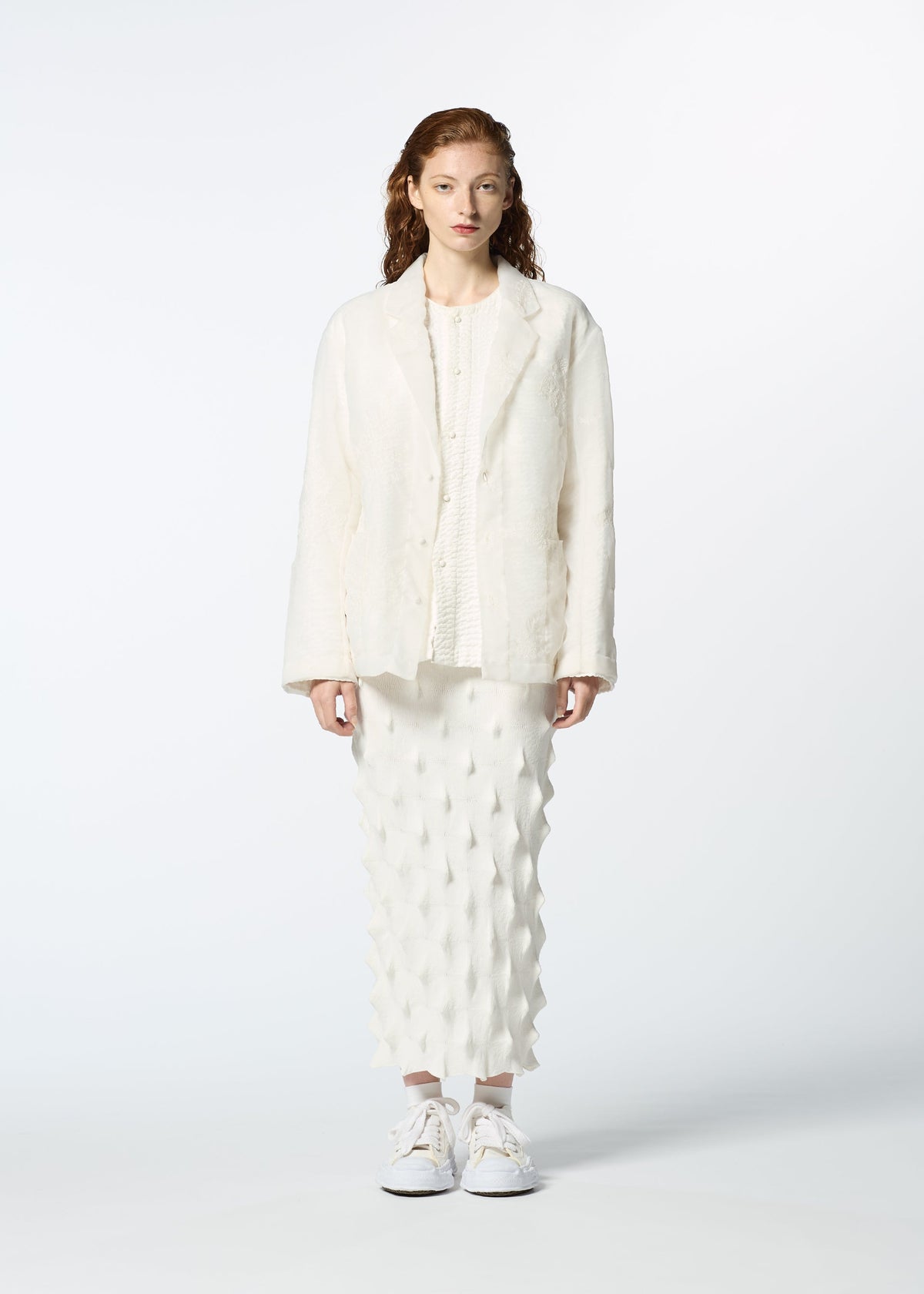 A person with wavy red hair faces forward, wearing HaaT's GLASSLEAF off-white Bhill-embroidered textured jacket and matching patterned midi skirt, paired with white sneakers, set against a plain white background. view 6 of 8