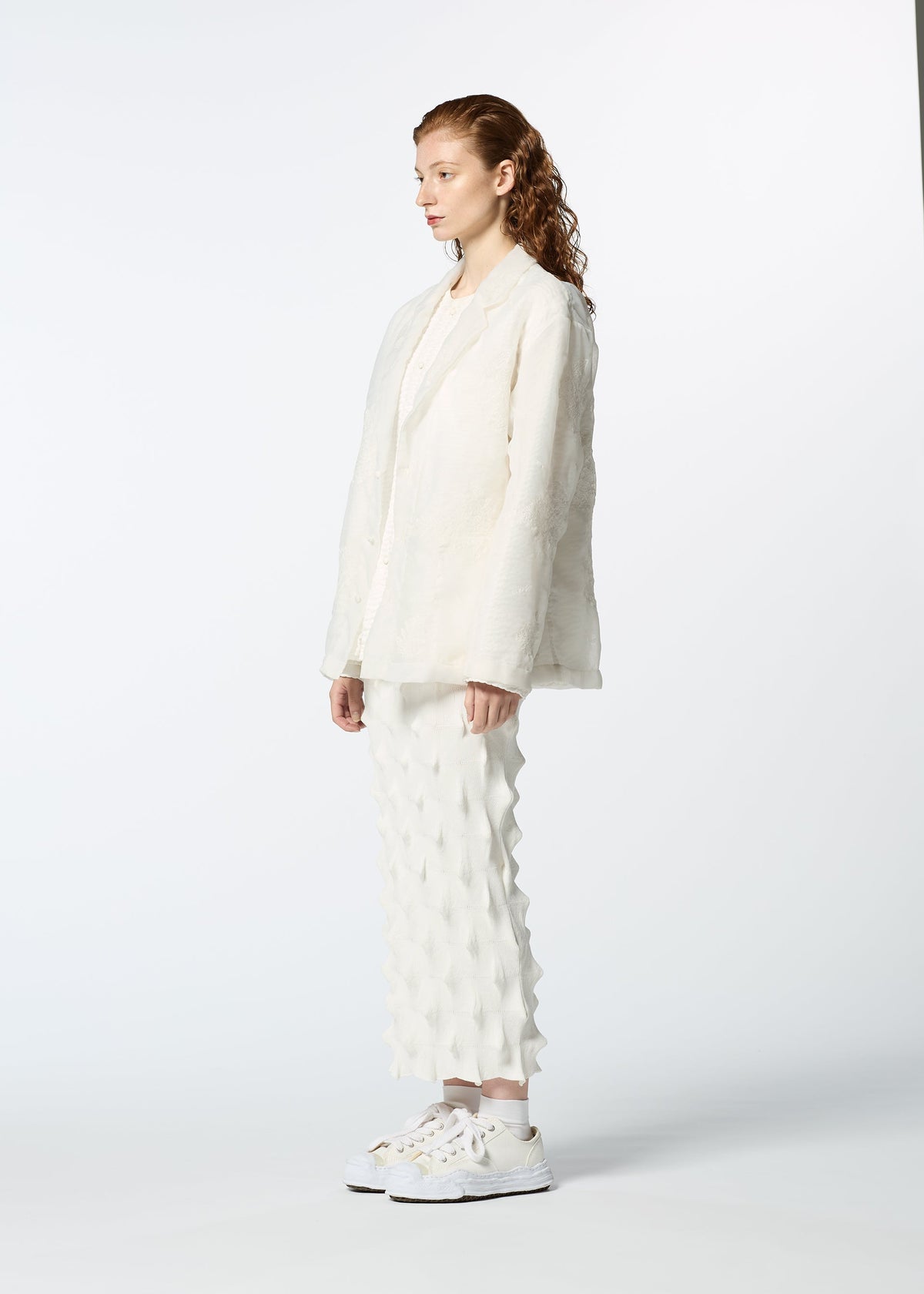 A person with long curly hair stands in profile wearing HaaT's GLASSLEAF off-white textured blazer with Bhill embroidery, matching pants, and white sneakers against a plain light background. view 7 of 8