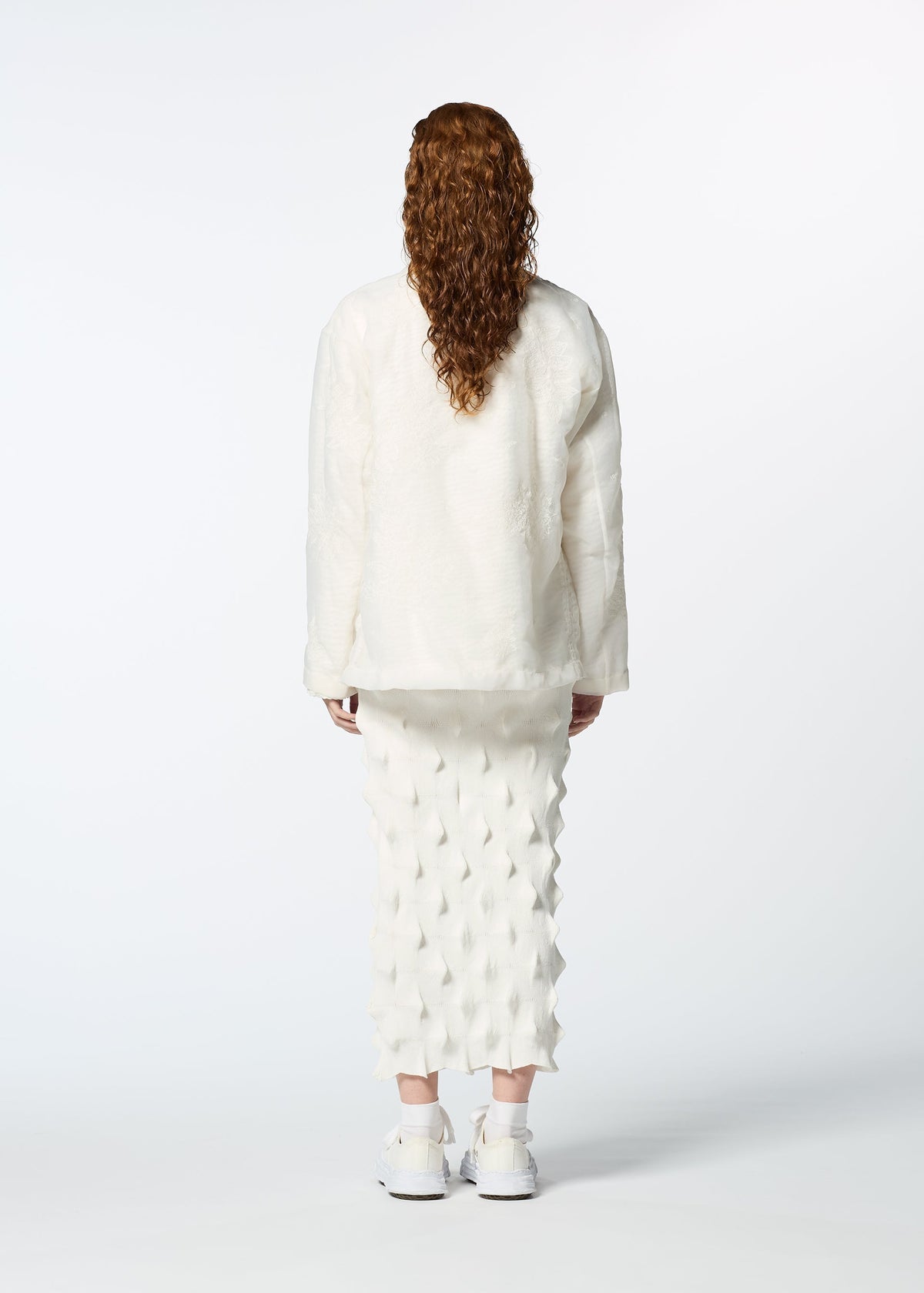 A person with long curly hair stands facing away, wearing HaaT’s GLASSLEAF textured white jacket and geometric patterned skirt with subtle Bhill embroidery, paired with white sneakers, against a plain white background. view 8 of 8