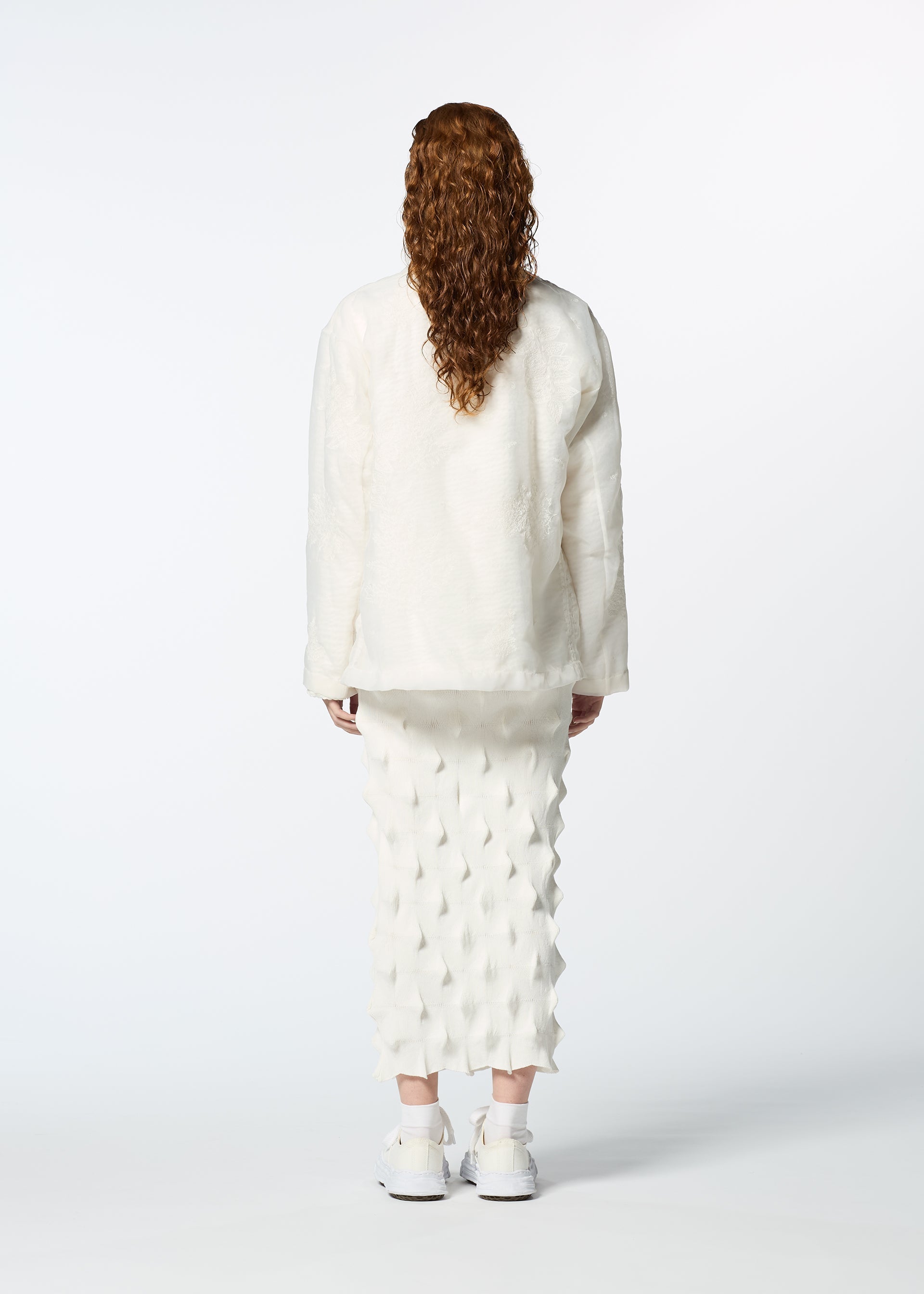 Zoomed view A person with long curly hair stands facing away, wearing HaaT’s GLASSLEAF textured white jacket and geometric patterned skirt with subtle Bhill embroidery, paired with white sneakers, against a plain white background.