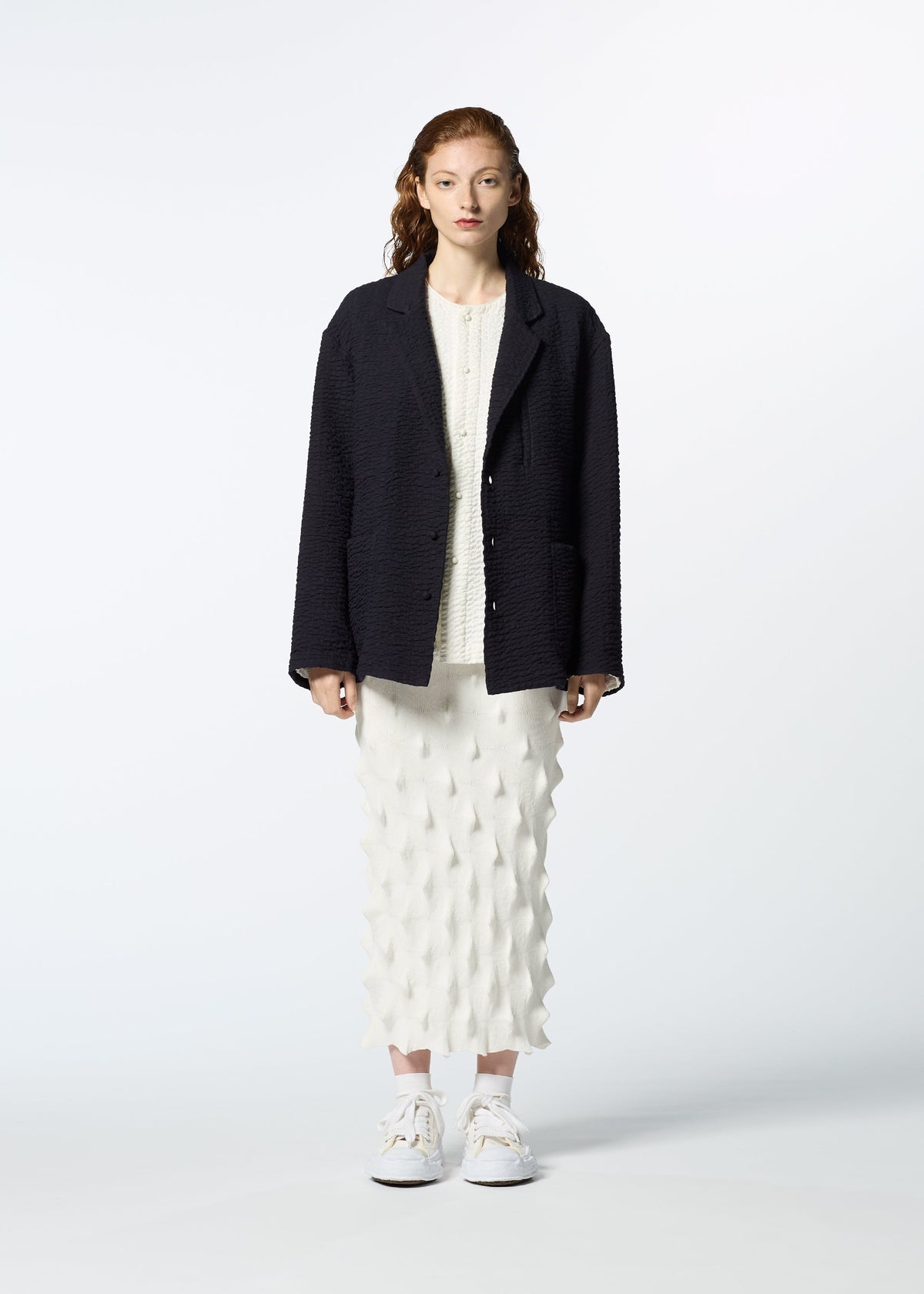 A woman with wavy brown hair faces forward in a HaaT 3 MUL MACHINE KABIRA '26SS textured black jacket over a white top and a mid-length white skirt with geometric pattern, paired with white sneakers, against a light background. view 4 of 8