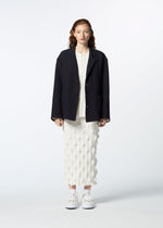 A woman with wavy brown hair faces forward in a HaaT 3 MUL MACHINE KABIRA '26SS textured black jacket over a white top and a mid-length white skirt with geometric pattern, paired with white sneakers, against a light background.