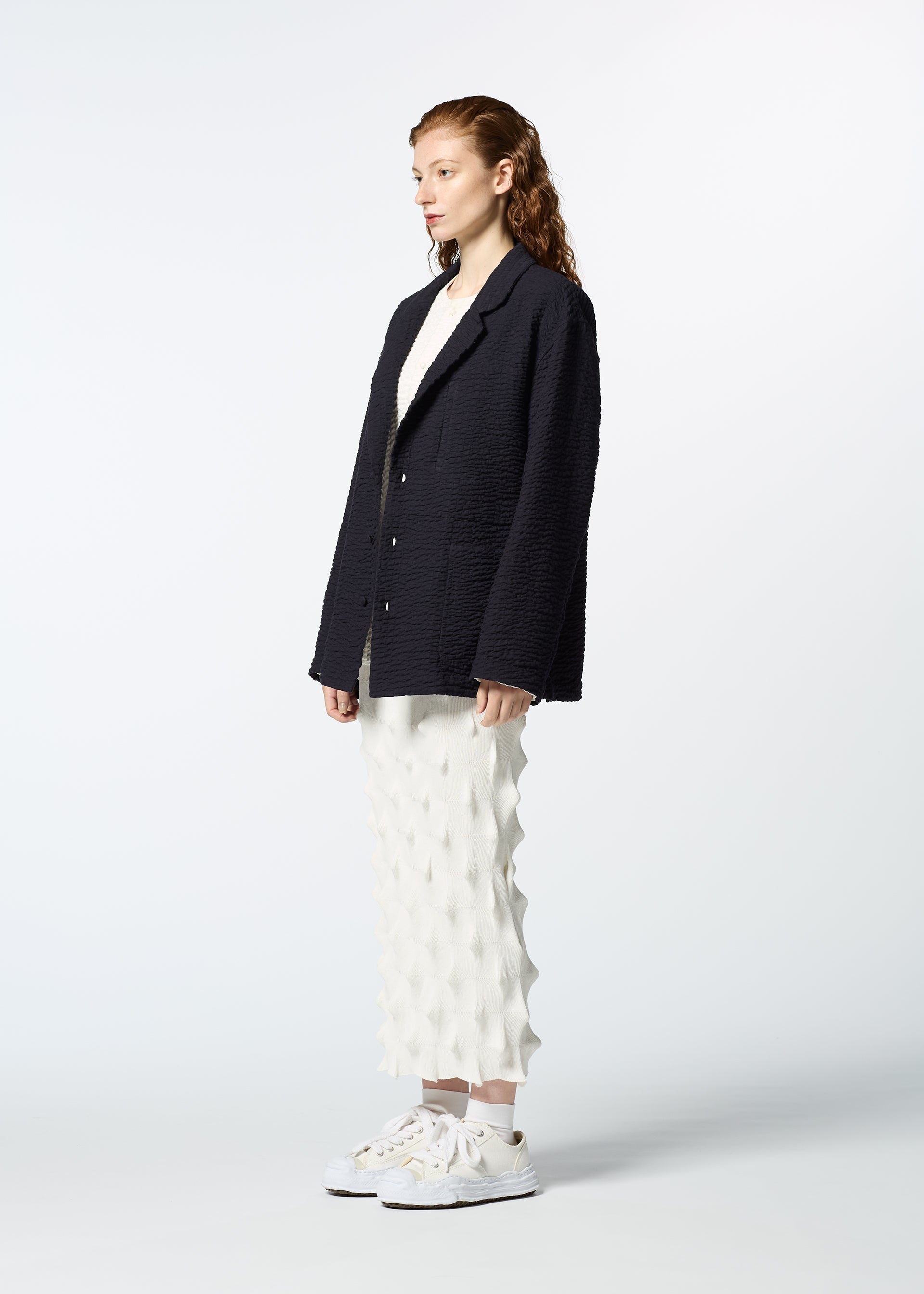 Zoomed view A woman in profile stands against a plain background, wearing HaaT's 3 MUL MACHINE KABIRA '26SS jacket with Indian embroidery over a white top and a unique geometric-patterned white skirt, paired with white sneakers.