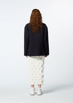 A person with long, curly hair faces away, wearing the HaaT 3 MUL MACHINE KABIRA '26SS textured black jacket, a white mid-length embroidered skirt, and white sneakers against a plain light background.