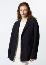 A person with long, wavy red hair wears HaaT's 3 MUL MACHINE KABIRA '26SS black textured jacket with Indian embroidery over a white shirt and pants, standing against a plain light background.