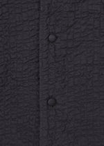 Close-up of the HaaT 3 MUL MACHINE KABIRA '26SS jacket in textured black with a crinkled pattern, vertical seam, and two fabric-covered round buttons, inspired by unisex silhouettes and subtle Indian embroidery.