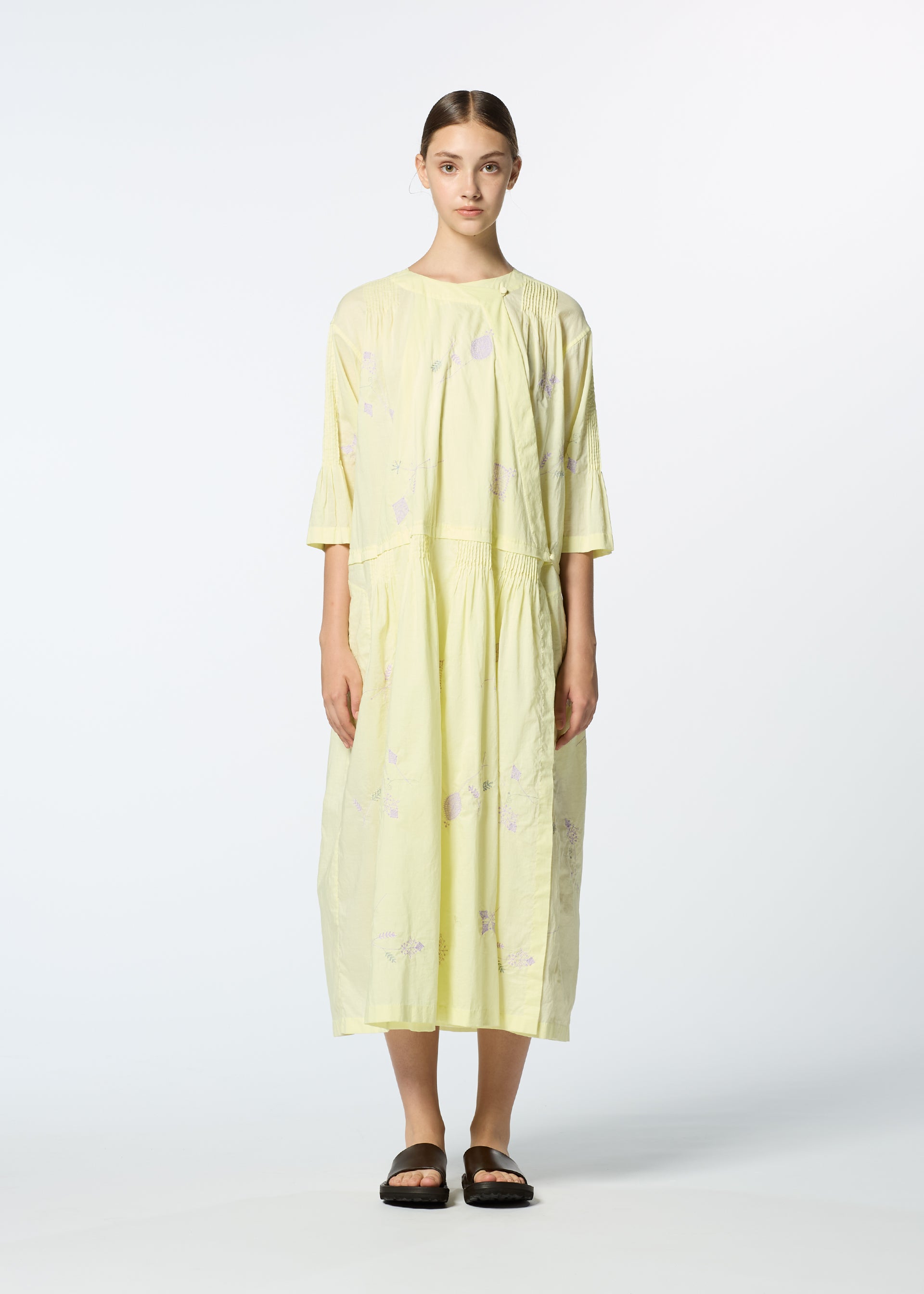 Zoomed view A woman stands against a plain background wearing HaaT’s DAMI LEAVES, a loose, light yellow handmade Indian dress with three-quarter sleeves and subtle floral patterns, paired with black flat sandals.