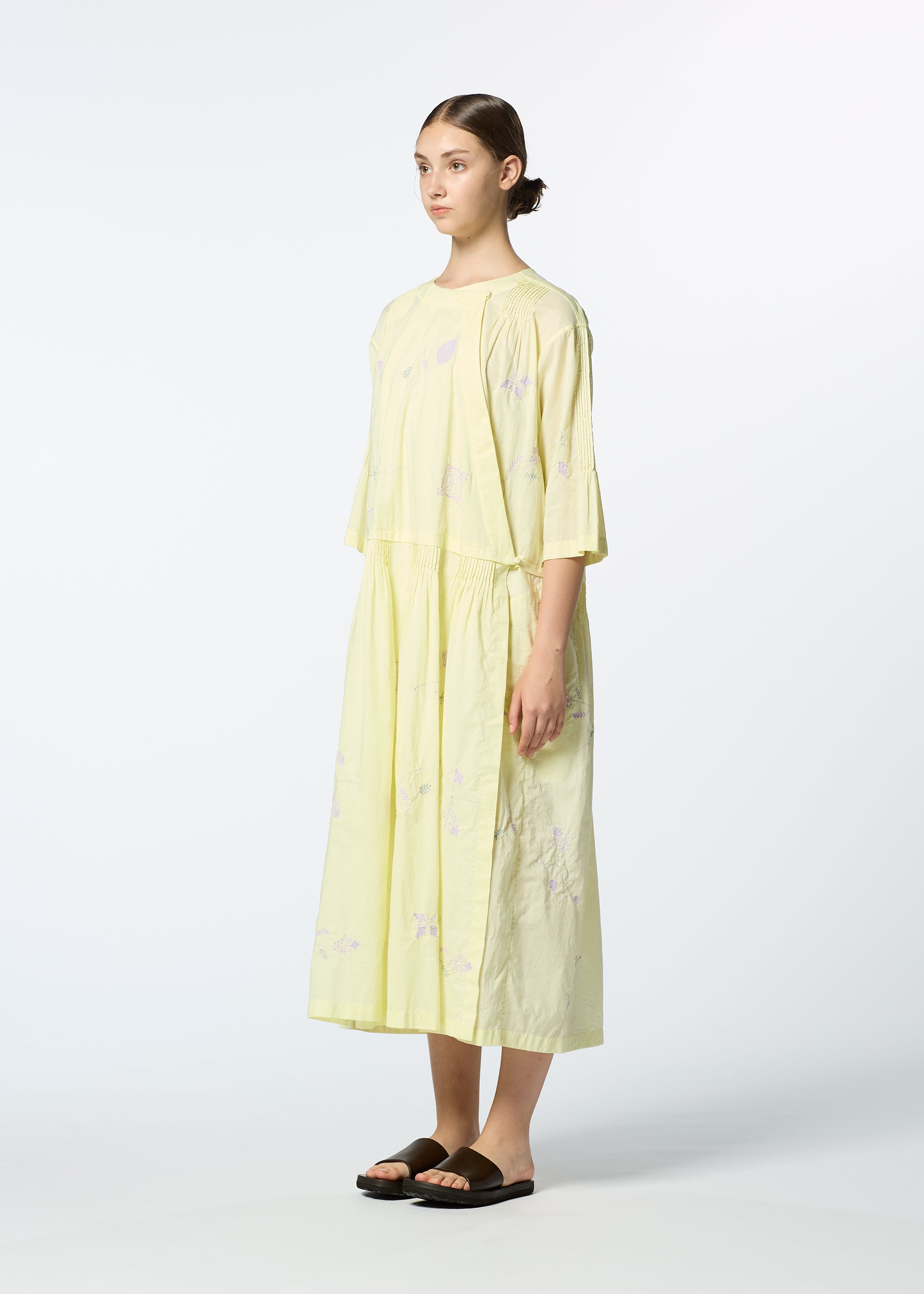 Zoomed view A woman in profile wears the DAMI LEAVES handmade Indian dress by HaaT—a loose, light yellow, mid-length piece with subtle Chikan embroidery—paired with black sandals, her hair tied back, set against a plain white background.