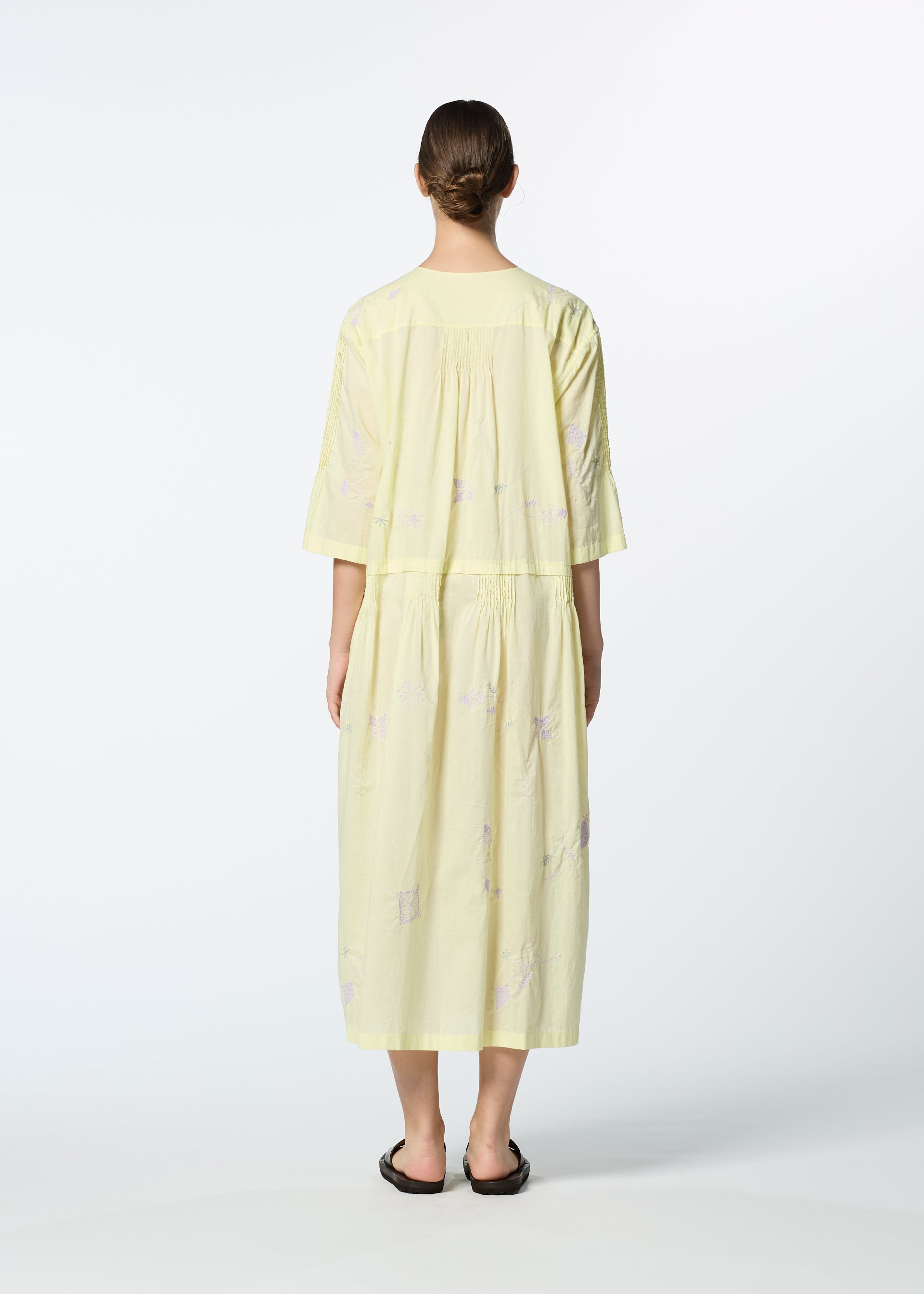 Zoomed view A person with dark hair in a low bun stands facing away, wearing HaaT’s DAMI LEAVES, a light yellow, loose-fitting handmade Indian dress with elbow-length sleeves and black sandals against a plain light background.