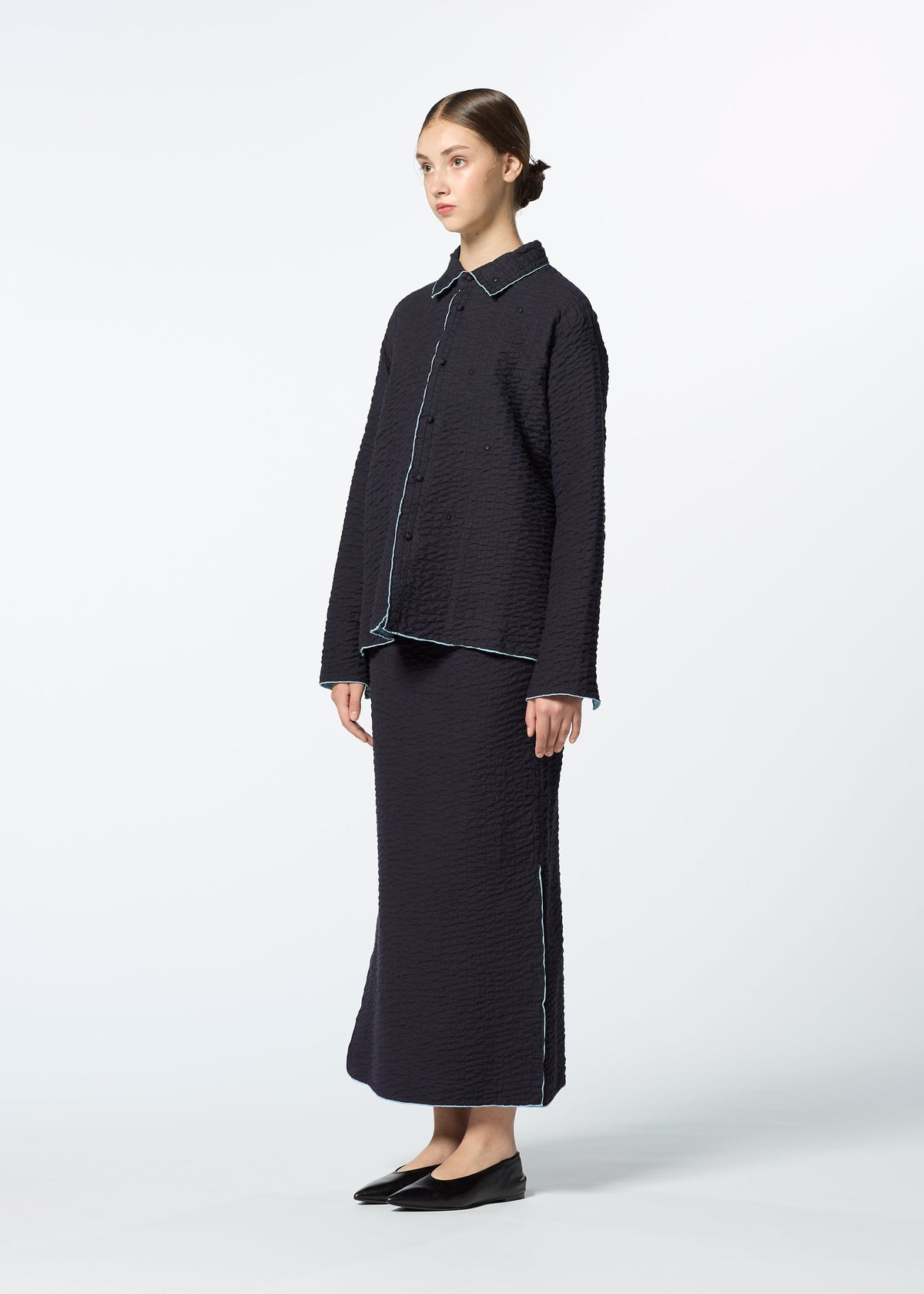 A woman stands against a plain background in HaaT’s DUAL LAYER: a textured, dark navy button-up shirt paired with a matching long skirt. Her black flats, tied-back hair, and handwoven cotton jacket complete the look. view 7 of 8