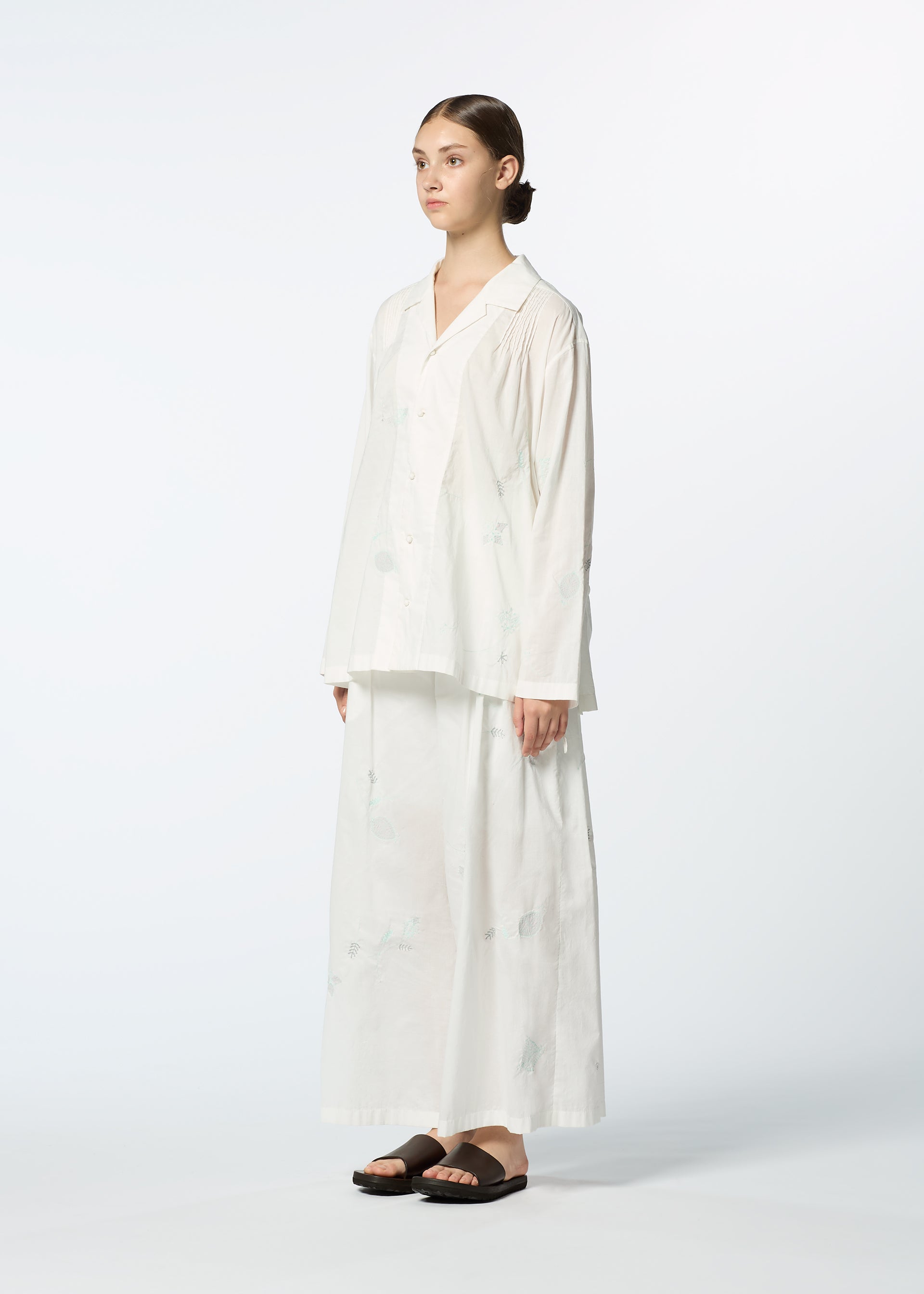 Zoomed view A person stands against a plain background wearing HaaT's DAMI LEAVES set: a loose, long-sleeve white shirt and matching long skirt with subtle Chikan embroidery. Their brown hair is pulled back, paired with black sandals.