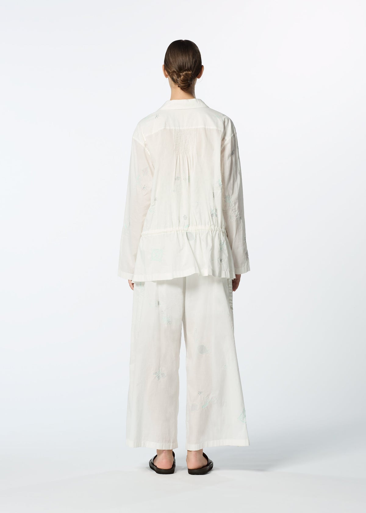 A person stands with their back to the camera, wearing HaaT's DAMI LEAVES loose white long-sleeve shirt with delicate Chikan embroidery and wide-leg pants, paired with dark slip-on sandals, set against a plain light background. view 9 of 10