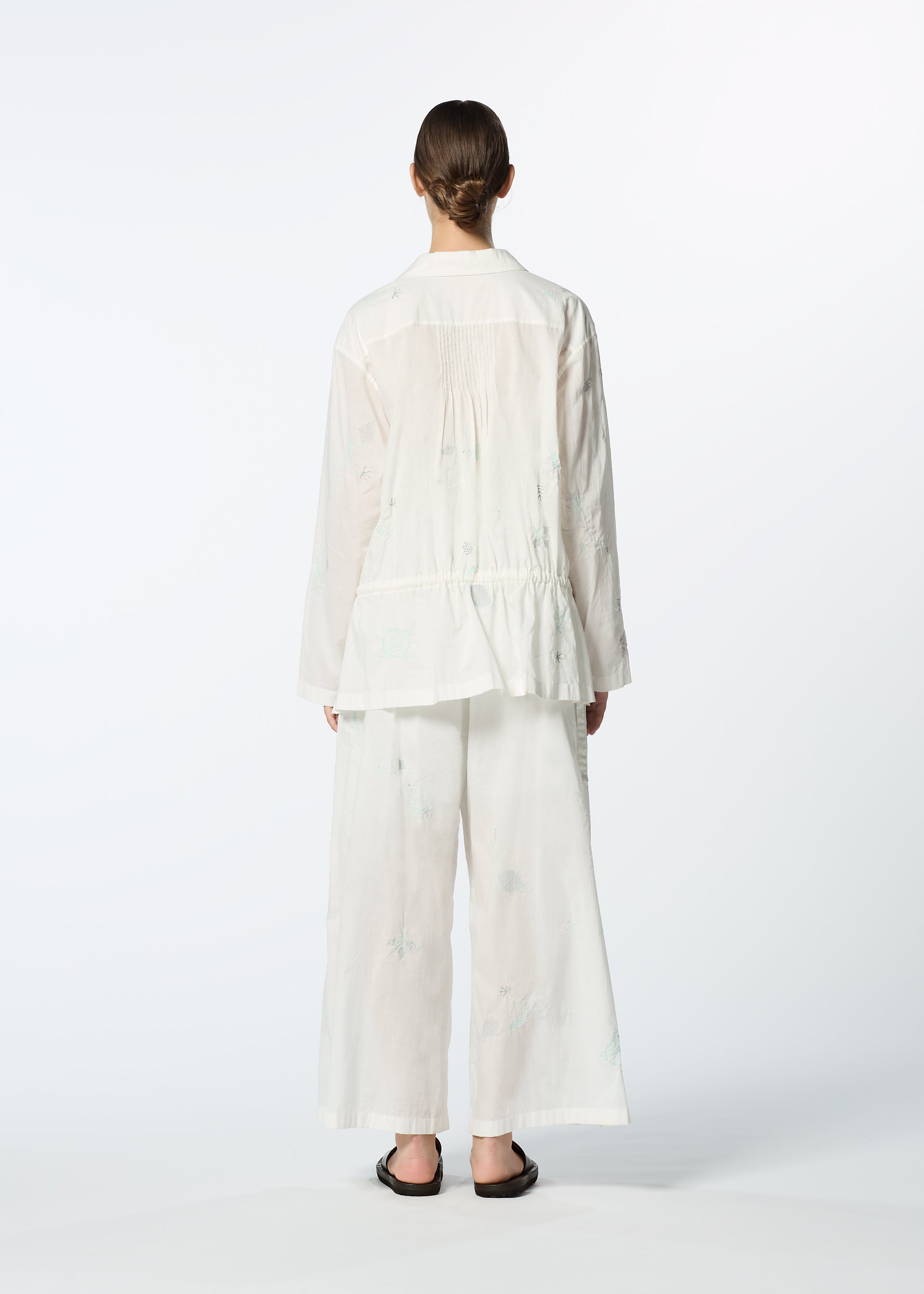 Zoomed view A person stands with their back to the camera, wearing HaaT's DAMI LEAVES loose white long-sleeve shirt with delicate Chikan embroidery and wide-leg pants, paired with dark slip-on sandals, set against a plain light background.