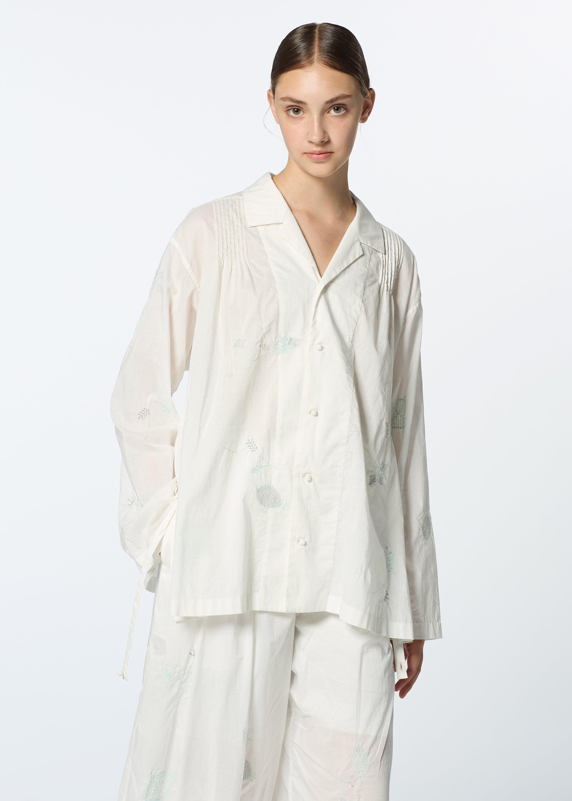 Zoomed view A young woman with brown hair tied back wears the DAMI LEAVES set by HaaT—a relaxed, pajama-style outfit with subtle Chikan embroidery and wide-leg pants, contrasting against a plain, light background.