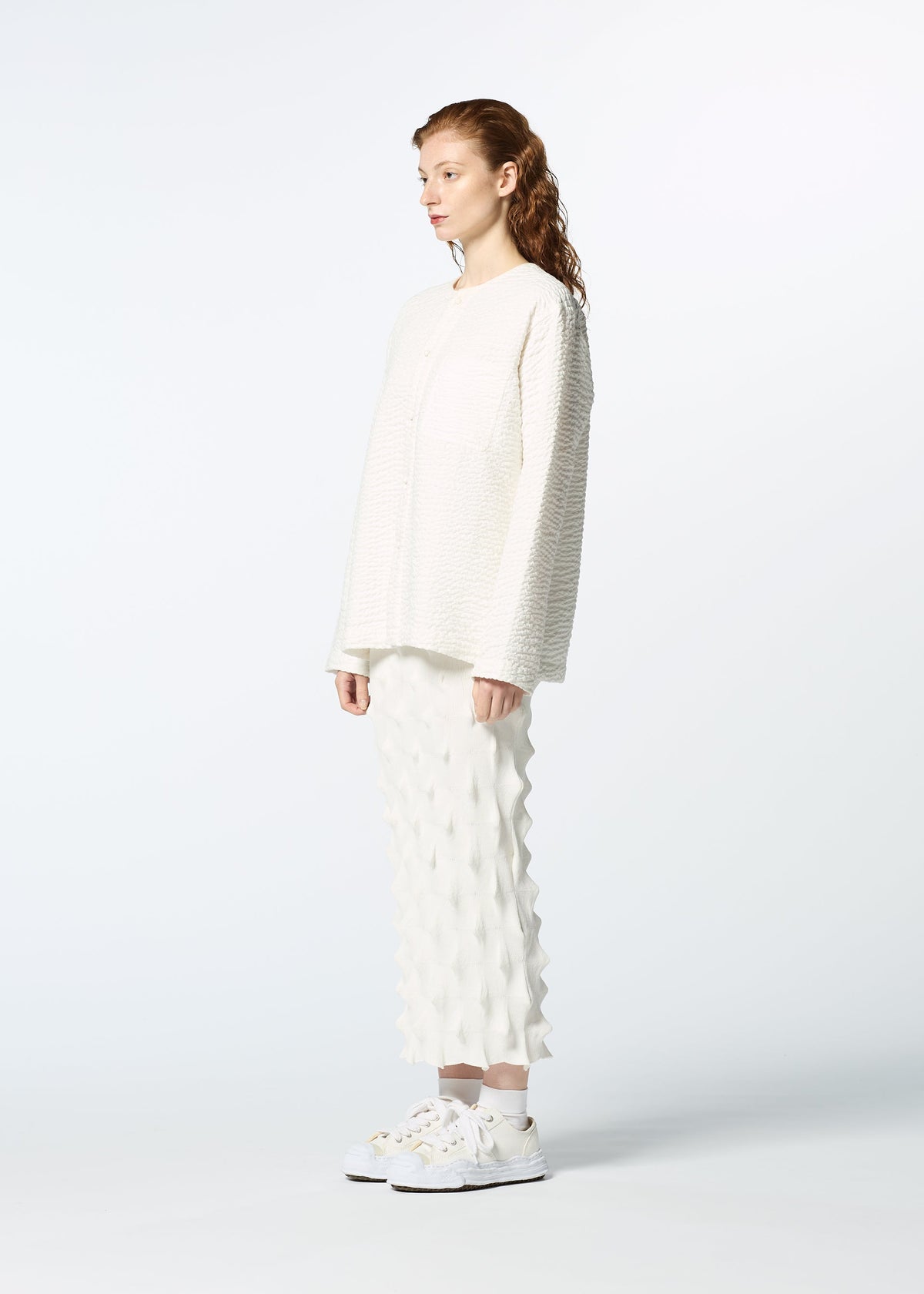 A woman wears the HaaT 3 MUL MACHINE KABIRA '26SS set—an off-white, collarless shirt and matching skirt with a 3D geometric pattern inspired by Indian embroidery—paired with white sneakers, standing left against a white background. view 7 of 9