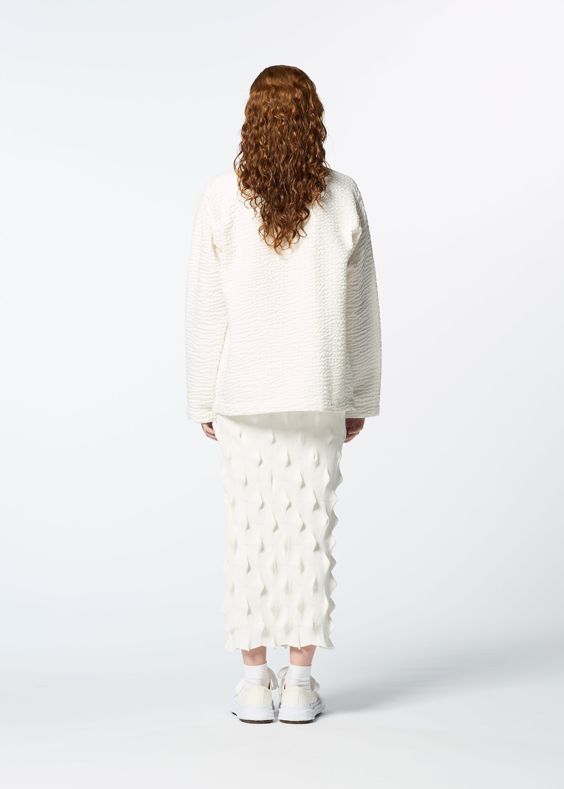 Zoomed view A person with long, curly brown hair faces away, wearing HaaT's 3 MUL MACHINE KABIRA '26SS—a collarless embroidered shirt, white textured skirt, and white sneakers—set against a plain white background.