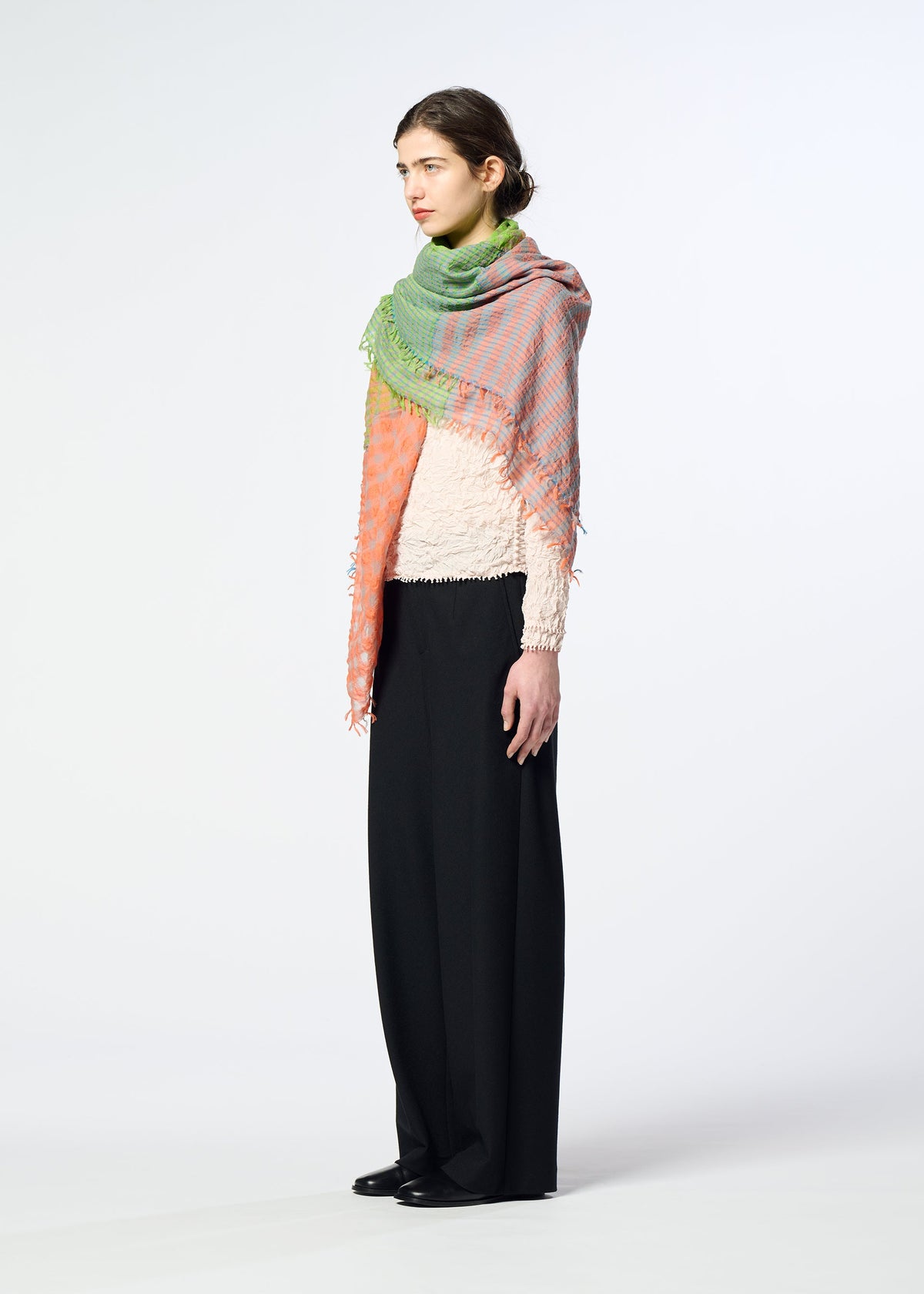 A woman stands sideways in a textured white top, wide black pants, and the HaaT TRIPLY SQUARE STOLE—silk-wool with hand-finished green and pink fringe—against a plain light background. view 5 of 6