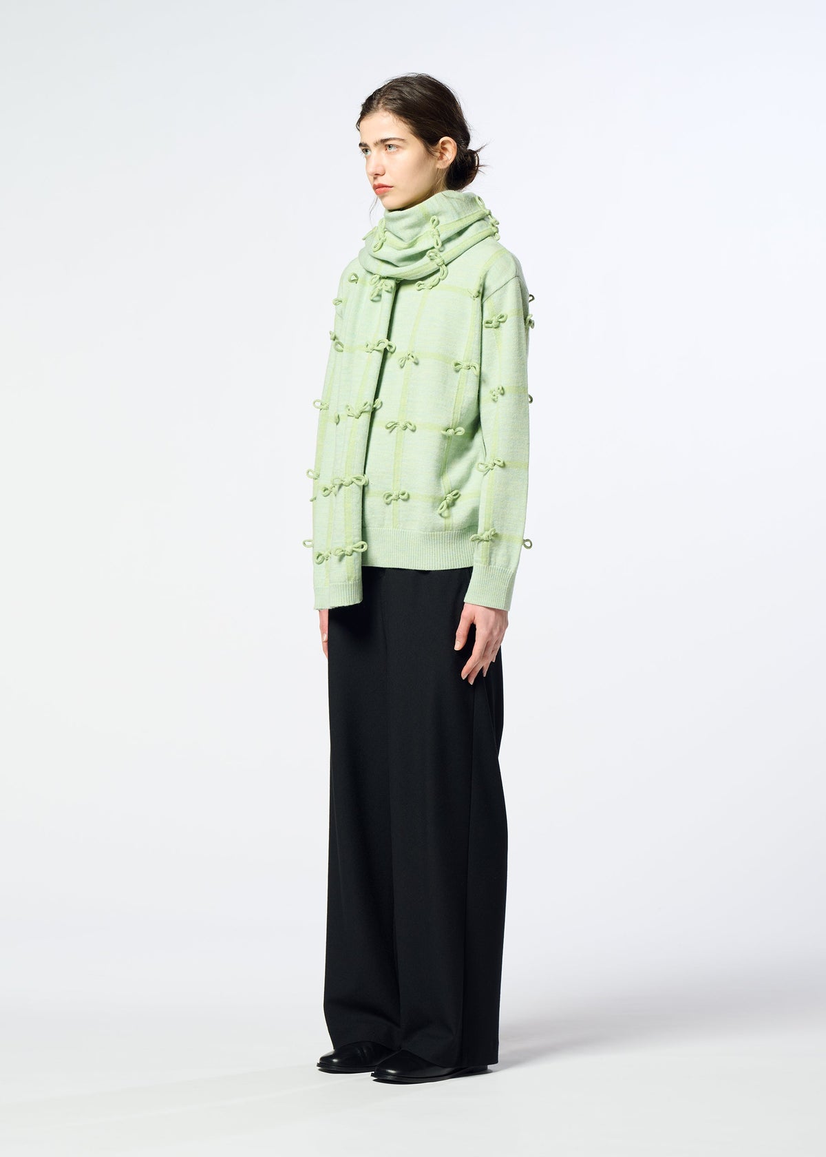 A person stands sideways against a light background, wearing HaaT’s RIBBON KNIT sweater in pale green with textured dots, a matching wool knit stole, wide black pants, and black shoes. view 6 of 7