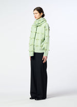 A person stands sideways against a light background, wearing HaaT’s RIBBON KNIT sweater in pale green with textured dots, a matching wool knit stole, wide black pants, and black shoes.