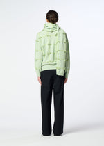 A person stands facing away, wearing HaaT’s RIBBON KNIT in light green with raised knot details, a hand-tied fulled wool knit stole, and loose black pants against a plain white background.