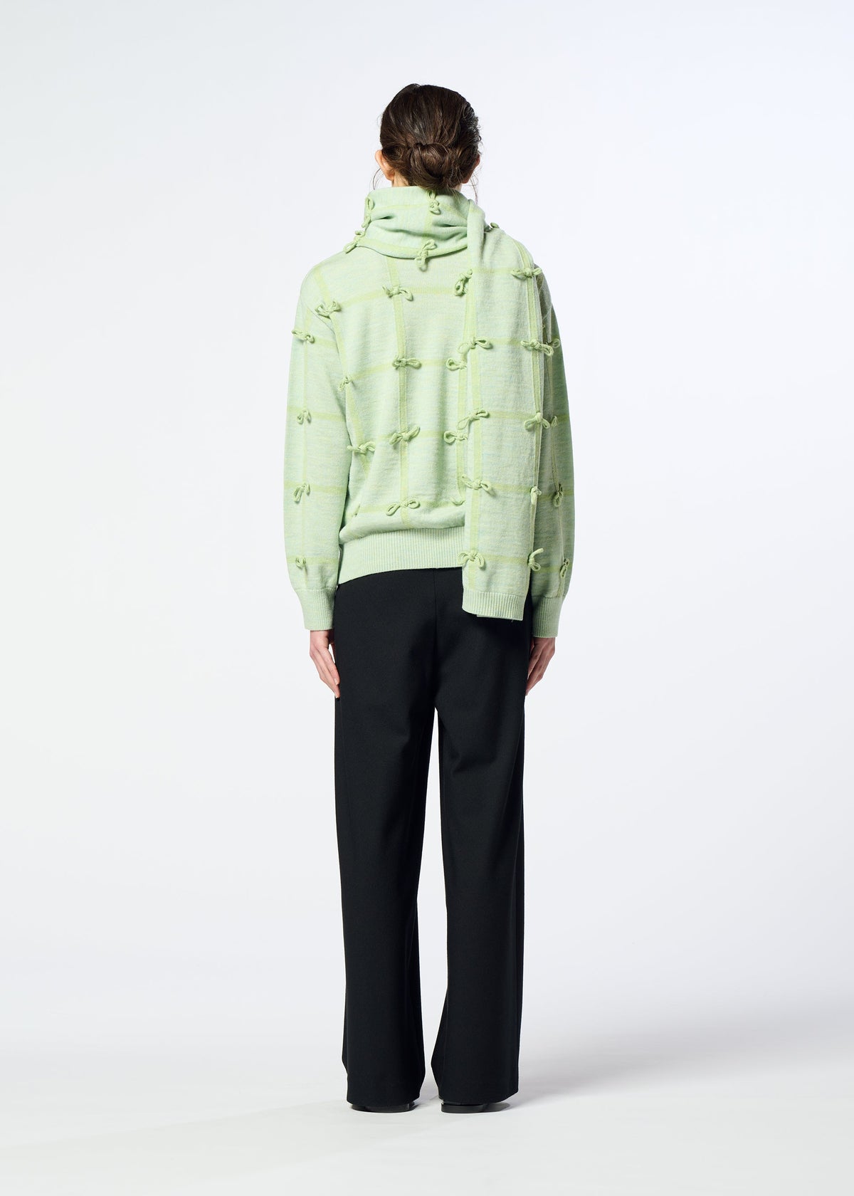 Zoomed view A person stands facing away, wearing HaaT’s RIBBON KNIT in light green with raised knot details, a hand-tied fulled wool knit stole, and loose black pants against a plain white background.
