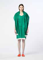 A person with straight brown hair stands against a plain white background, wearing HaaT’s WALL WHITE HEM, a loose bright green sleeveless dress with white trim, orange shoes, and a delicate chiffon stole with a subtle Nassen print.