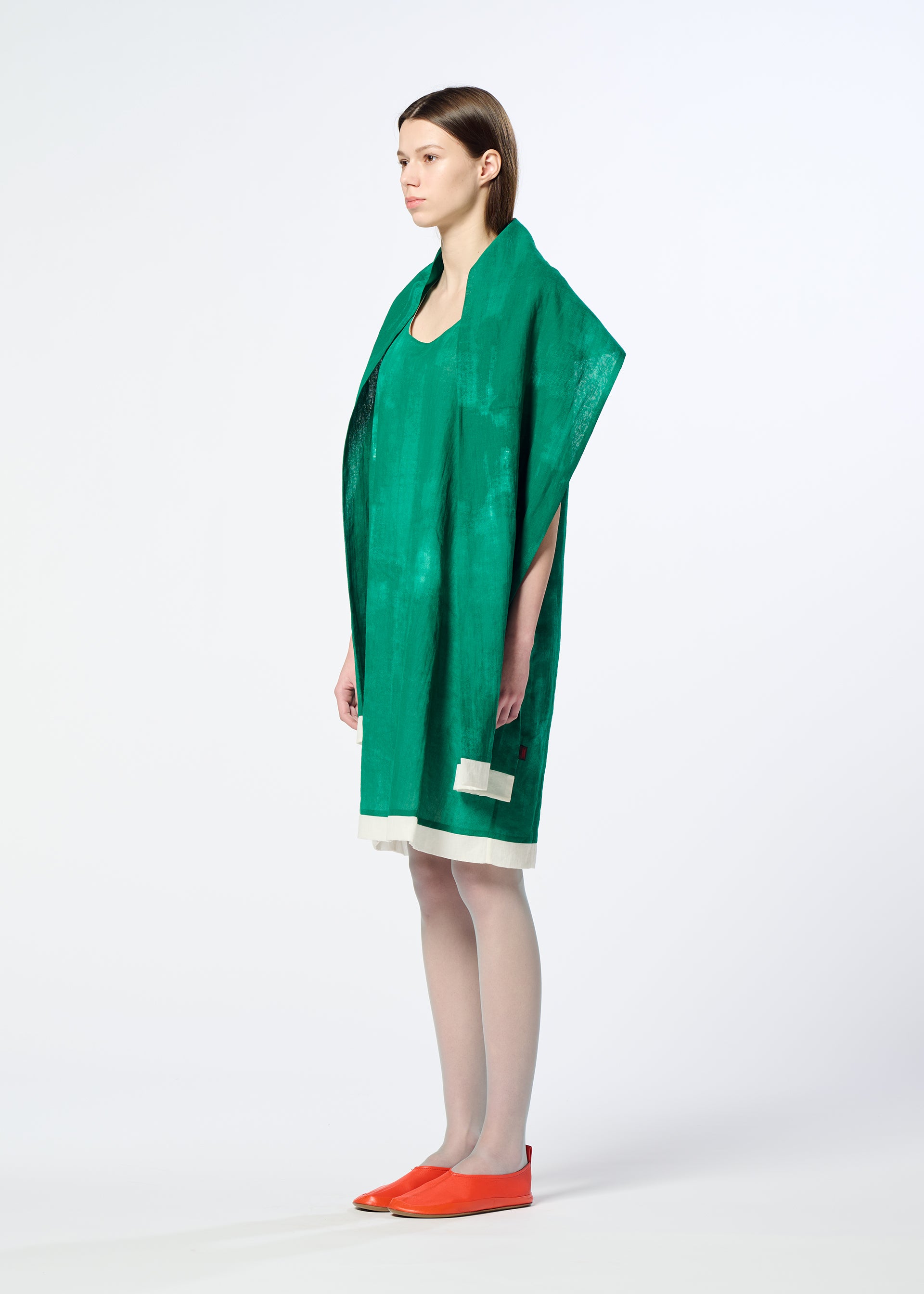 Zoomed view A woman stands sideways wearing the HaaT WALL WHITE HEM, a loose green sleeveless dress with a wide collar and white-trimmed hem, styled with orange slip-ons and a chiffon stole against a plain white background.