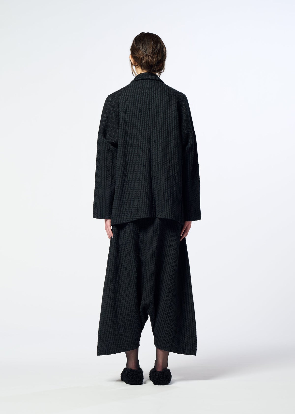 Zoomed view A person stands facing away, wearing a loose-fitting, textured black SAKIORI jacket by HaaT with wide cropped pants and textured black shoes, set against a plain white background.