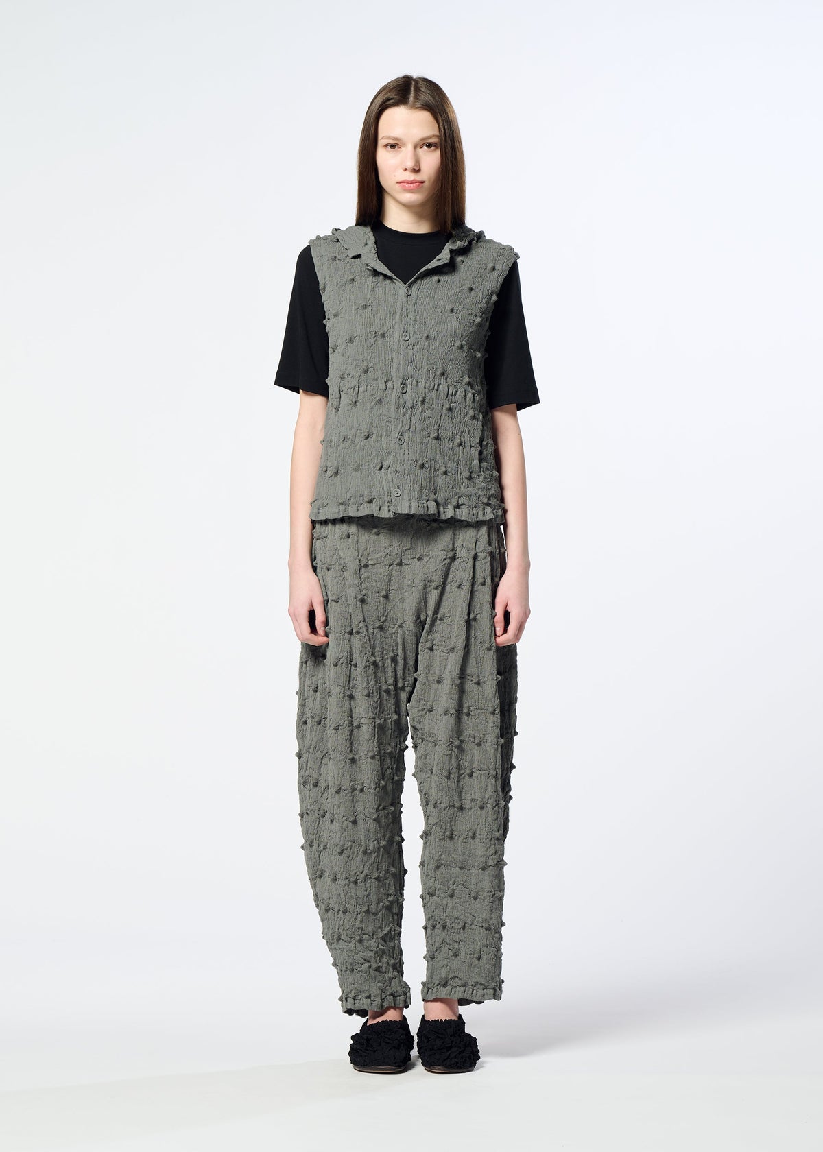 A model stands facing forward against a plain white background, wearing the HaaT KYO CHIJIMI SQUARE EMB set—a textured gray vest featuring embroidered square patterns, paired with matching pants over a black shirt. They have straight brown hair and are wearing black shoes. view 7 of 10