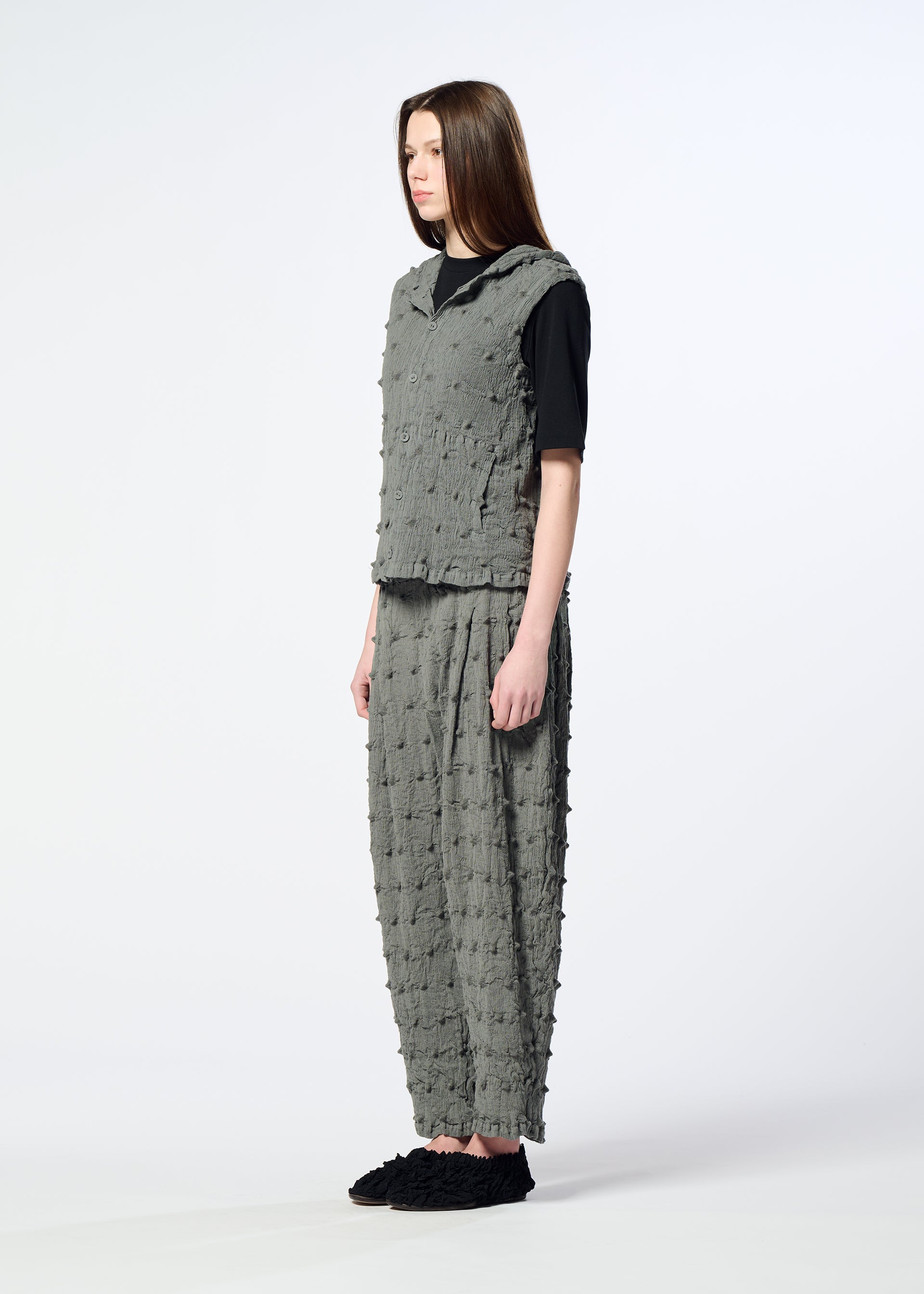Zoomed view A woman stands in profile against a plain white background, wearing the HaaT KYO CHIJIMI SQUARE EMB—a textured, loose-fitting gray top and matching pants—layered over a black shirt and paired with black slip-on shoes.