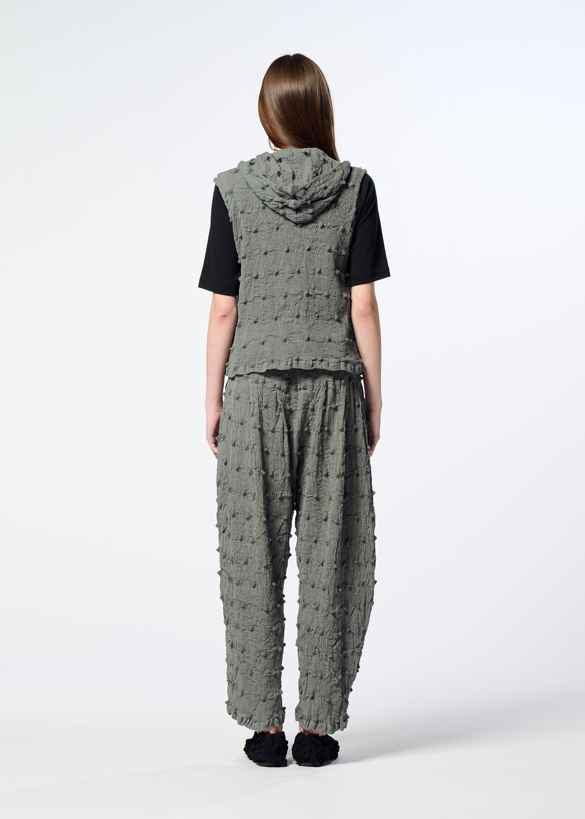 Zoomed view A person stands with their back to the camera, wearing the HaaT KYO CHIJIMI SQUARE EMB textured gray hooded vest and matching loose pants over a black shirt, set against a plain white background.
